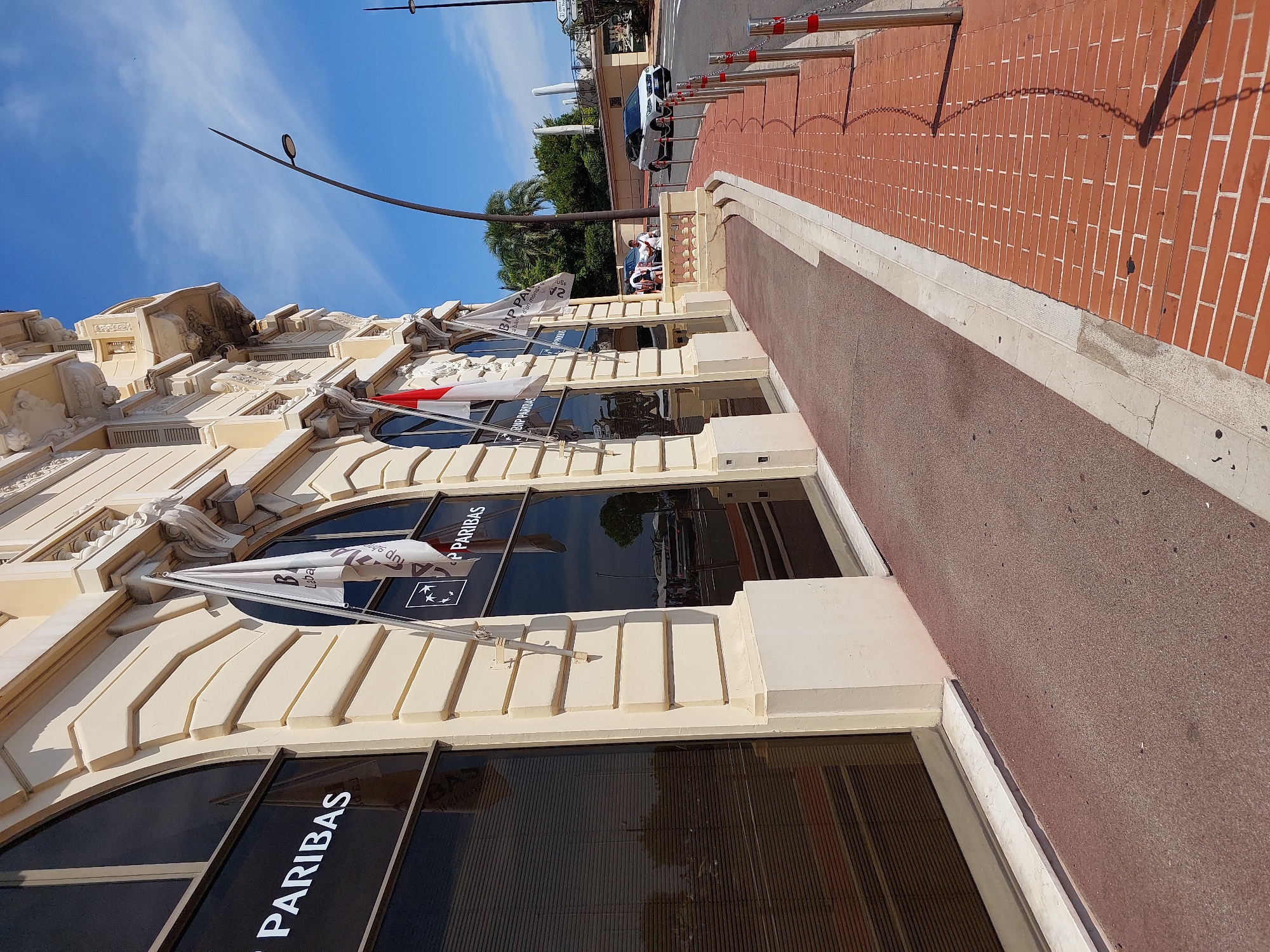 The image shows the exterior of the BNP Paribas bank on a sunny day in Monaco. The bank building is light beige with arched windows and decorative architectural elements. The name "PARIBAS" is visible on the windows in large white lettering. Two flags are displayed on the building, one white with a red stripe and another white with a red cross, likely representing Monaco. A sidewalk made of red bricks runs along the front of the building, bordered by a darker red pathway. Metal bollards with chains are installed along the edge of the sidewalk, separating it from the street. A white car and a darker car are visible on the road, with a lamppost arching over the street in the background. Trees and lush vegetation are visible behind a wall further down the street.