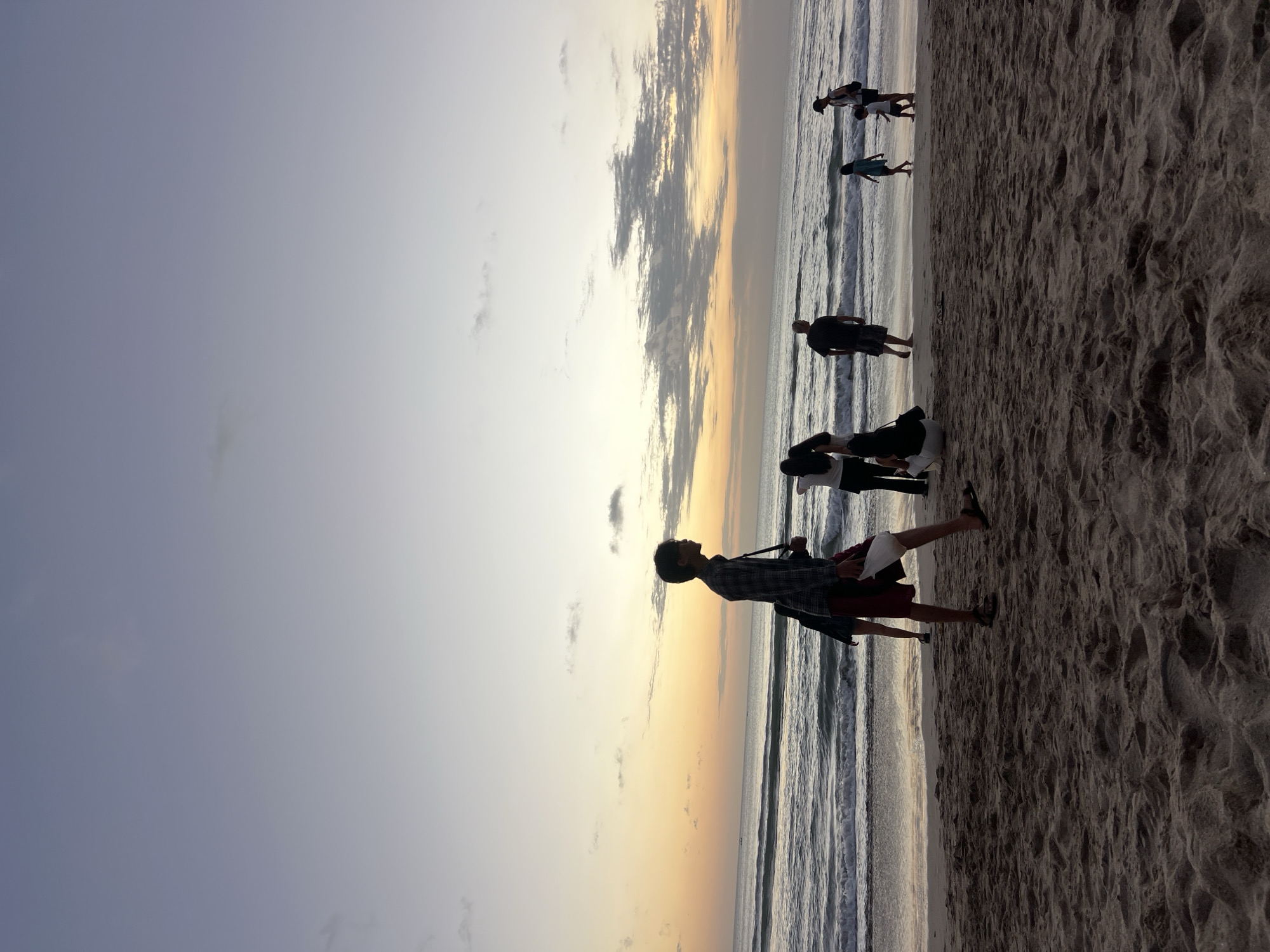 The image shows a beach scene at dusk in Kuta, Indonesia. The sky is a gradient of pale blue to yellow and orange near the horizon, with scattered clouds. Waves are rolling onto the sandy shore. Several people are visible on the beach. In the foreground, a person in a plaid shirt and red shorts walks from left to right, carrying a white bag. To their left and further back, another person is partially visible. In the middle ground, a group of people are standing and sitting, including a person in a black shirt and shorts and a child in a blue dress. Further to the right, another group includes a person in a black shirt and cap and two children. The sand is covered in footprints and textured from the tide. The overall impression is a tranquil evening on a popular Indonesian beach.