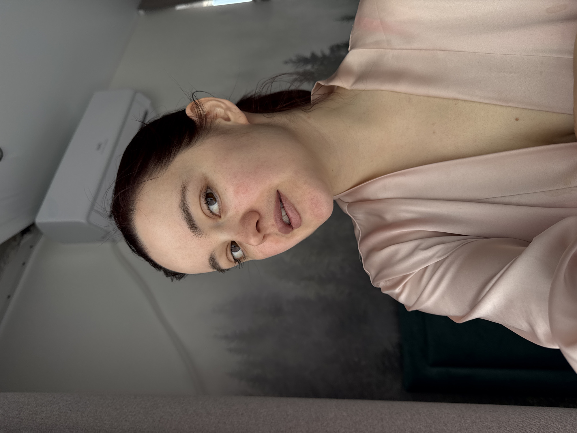 A medium close-up shot depicts a woman with dark hair pulled back, fair skin, and light-colored eyes. She is wearing a light pink, silky garment, possibly a robe or pajamas. Her head is slightly tilted to the right.
The background includes a light gray wall, a white air conditioning unit mounted high on the wall, and a wallpaper or mural depicting a dark forest scene. Part of a dark green object, possibly a chair or cushion, is visible in the lower right corner. The scene appears to be indoors.
The photograph was not taken in Izhevsk, Russia. There is no verifiable evidence to link the image to this location.