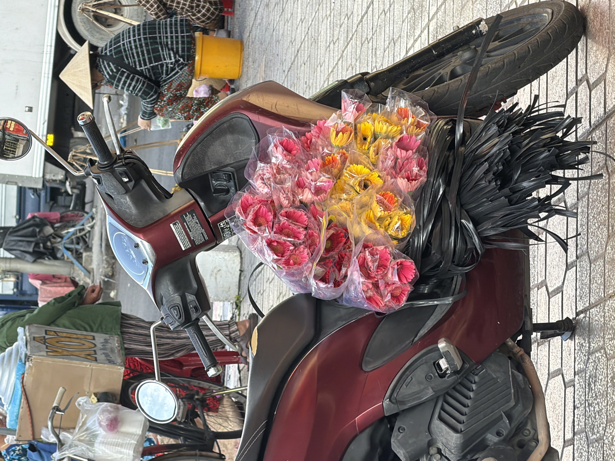 The image depicts a street scene in Nha Trang, Vietnam, focused on a red scooter with a bouquet of pink and yellow gerbera daisies tied to the seat. The flowers are packaged in clear plastic, with a bundle of black plastic ties beneath them. In the background, a person wearing a conical hat and a plaid shirt sits on a yellow bucket, seemingly preparing goods. Another person in a green jacket stands near a cart with a box labeled "XÔI," indicating a food vendor. The ground is paved with hexagonal tiles, and a portion of a truck and other market elements are visible. The overall scene suggests a bustling outdoor market environment.