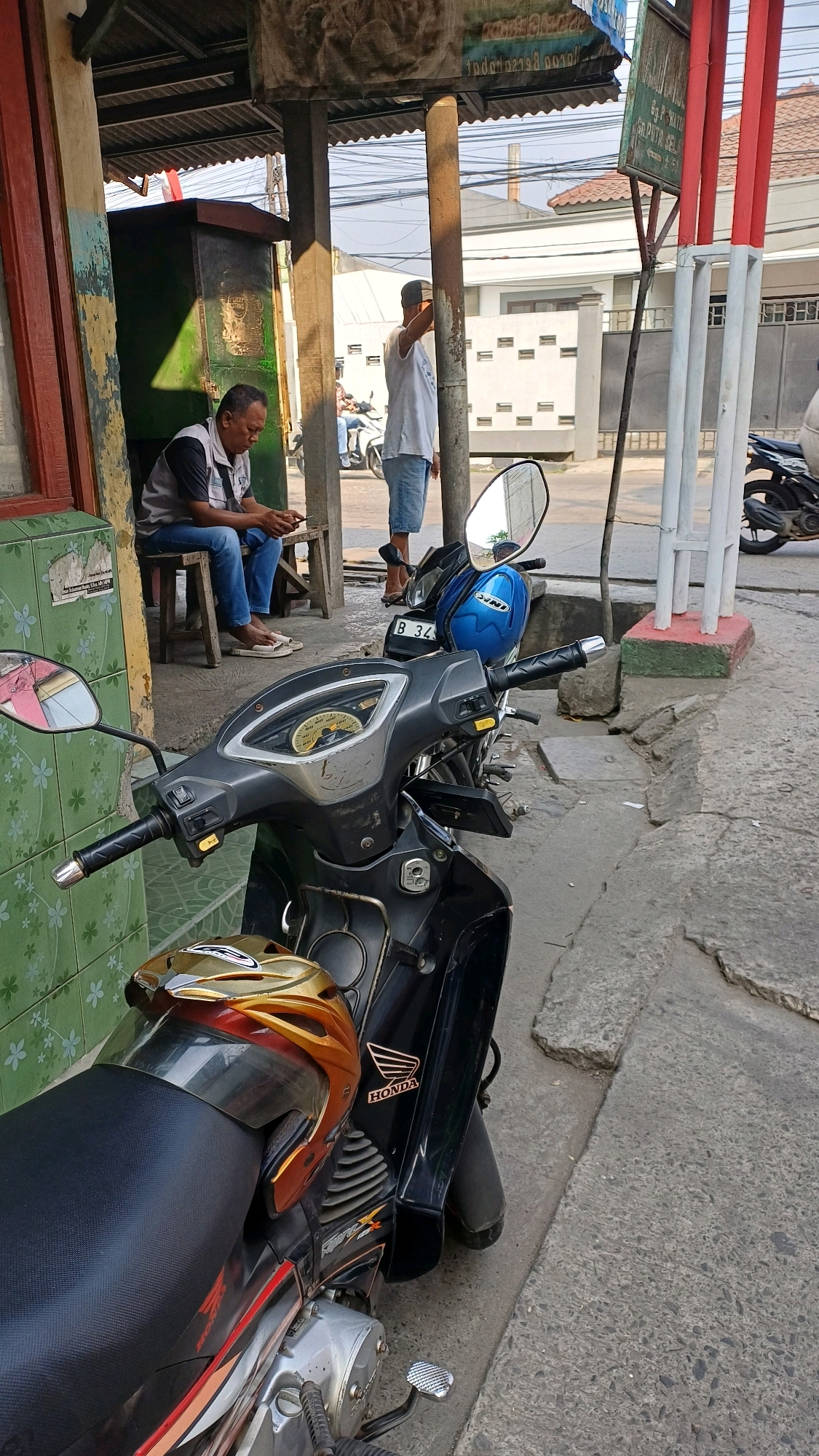 In Gunung Putri, Indonesia, a man sits on a wooden stool in the shade of a covered structure, looking at his mobile phone. He is wearing a dark vest over a light-colored shirt and blue jeans. In front of him, to the right, a black motorcycle is parked, its front fairing and handlebars prominently visible. A blue helmet rests on the seat of the motorcycle, and a gold and red helmet is placed on the rear of the seat. Another motorcycle, partially visible, is parked further down the street, to the right of the first. A white pillar and a red and white striped pole stand near the motorcycles. The environment appears to be an urban or semi-urban setting with buildings and a street visible in the background.