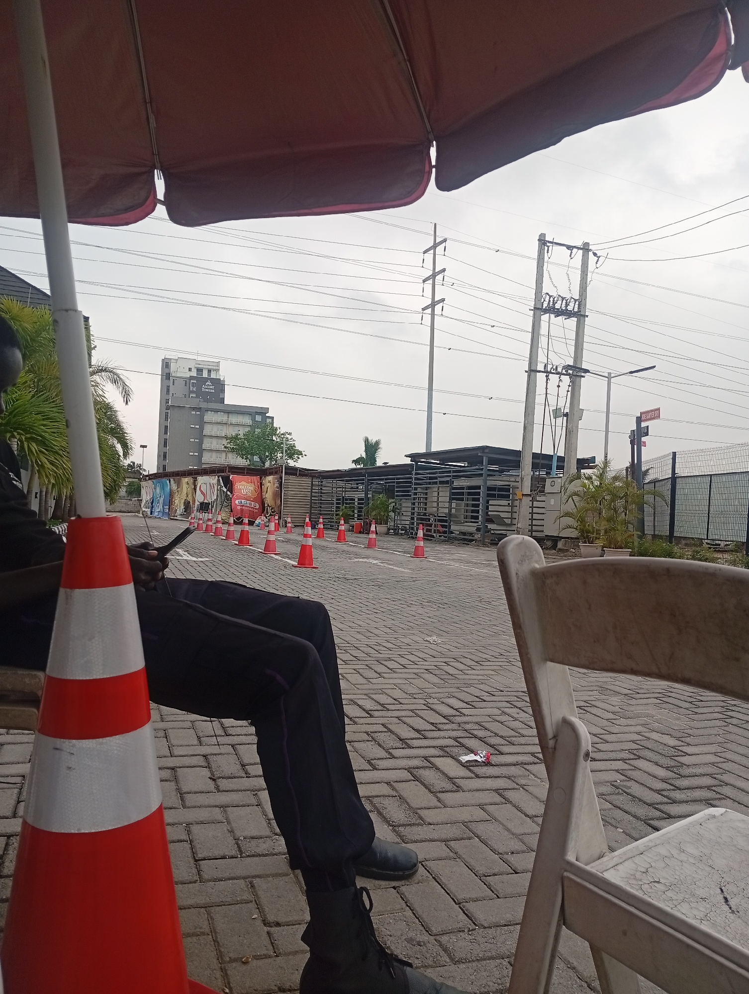 A person, partially obscured by a traffic cone, sits on a bench in Ilado, Nigeria. The person is wearing dark clothing and boots. In the background, a tall building with a sign that reads "ASCORT TOWER" stands against a cloudy sky. A row of orange traffic cones lines a paved area. Electrical poles and wires are visible in the distance, along with a fence. A white plastic chair is positioned in the foreground.