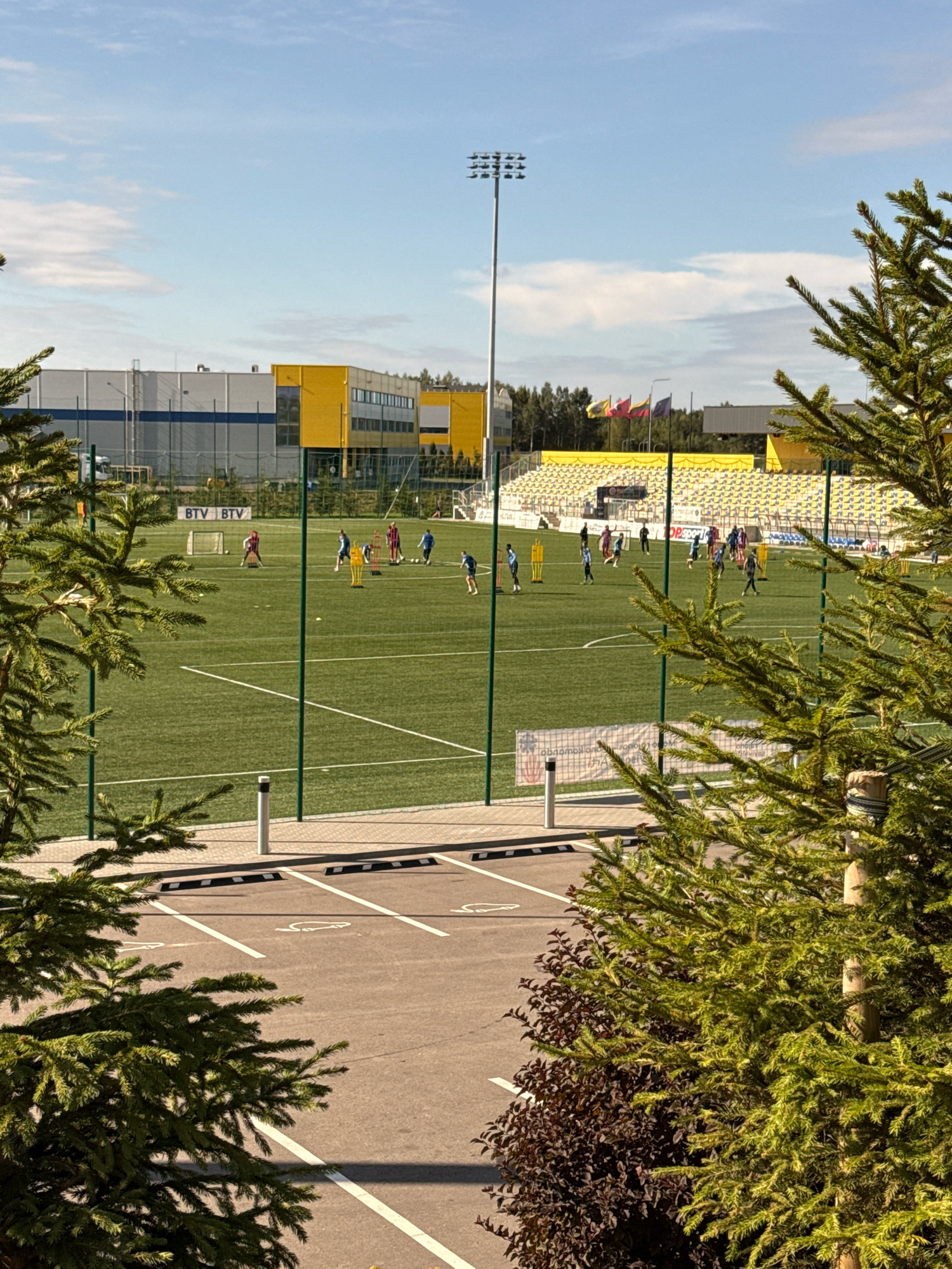 The image shows a football field with several people practicing. The field is green with white markings, and there are goalposts on either side. In the background, there are yellow and white stadium seats, a tall light pole, and some buildings. The sky is blue with some clouds. The scene appears to be set in a stadium or sports complex. The image is taken from a slightly elevated angle, with some greenery in the foreground, partially obscuring the view of the field. The people on the field are dressed in athletic wear, and some are kicking a ball or running. The overall impression is of a sunny day with an active sports event taking place.