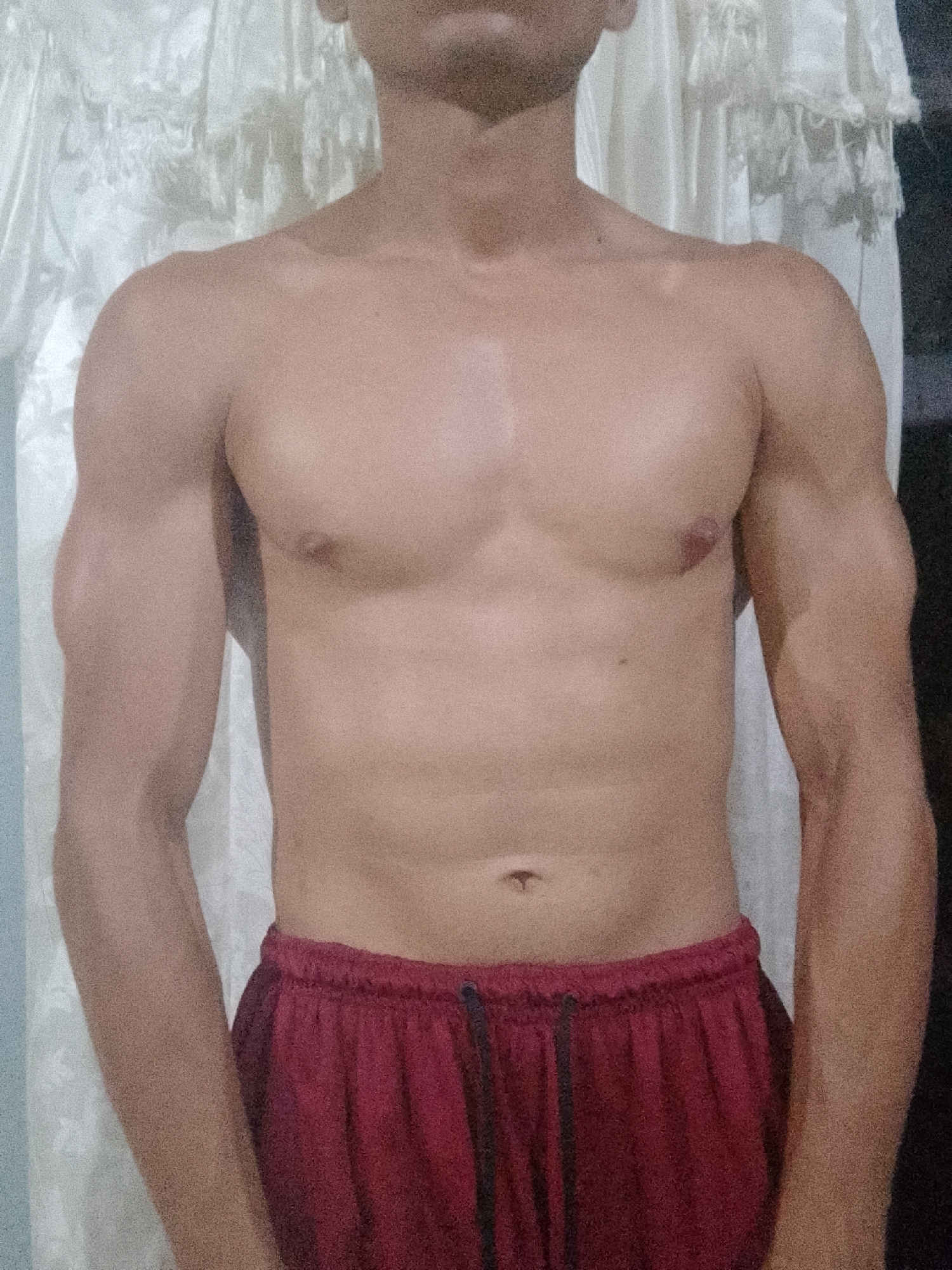 A shirtless individual with a visible physique stands in front of a white curtain, wearing red shorts with a drawstring. The upper portion of the body is visible, showcasing shoulders, chest, arms, and the upper abdomen. The background consists of a blurred, light-colored drape. There is no information in the image to indicate the location is Juan Griego, Venezuela.