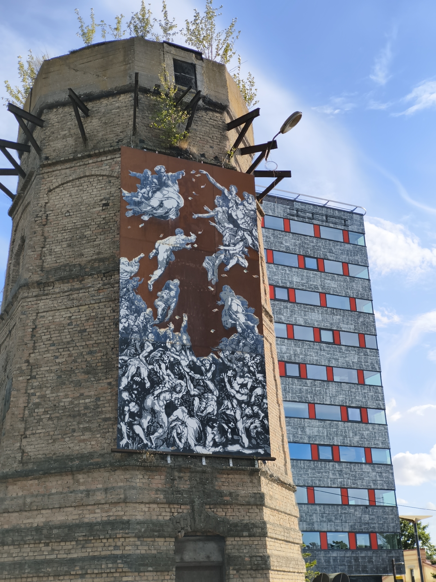The image captures a mural on the side of a cylindrical brick tower in Tallinn, Estonia. The mural depicts a scene with numerous figures, some appearing to be in motion or falling, rendered in black and white against a rust-colored background. Above the mural, the top of the brick tower shows signs of wear, with concrete and metal reinforcements, and vegetation growing from its upper edge. To the right of the tower, a modern, tall building with a grey facade and red-framed windows is visible against a bright blue sky with scattered white clouds. A streetlamp is mounted on the side of the brick tower, pointing towards the modern building.
