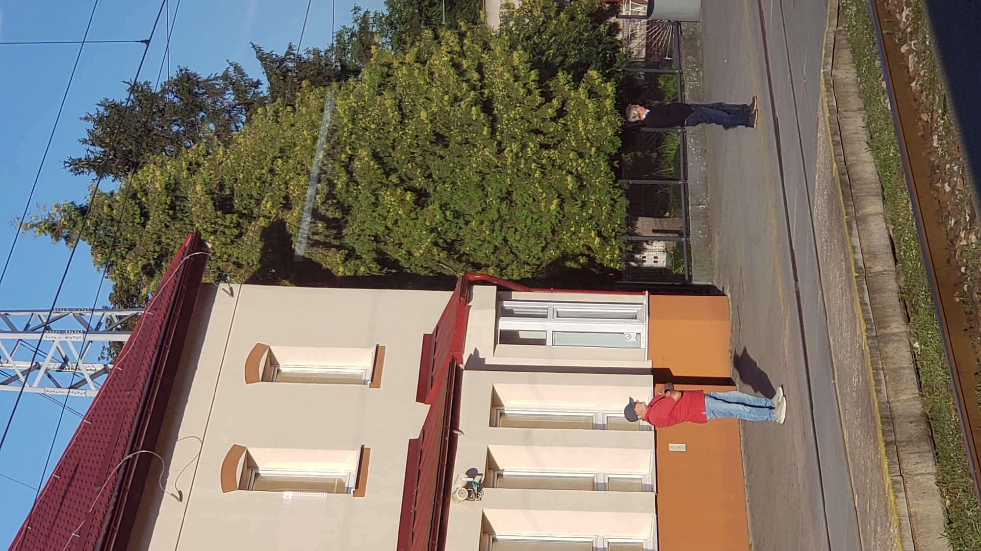 The image shows a train station platform in Spišská Nová Ves, Slovakia. Two men are standing on the platform. The man on the left is wearing a red polo shirt, blue jeans, and a blue baseball cap. He is standing with his arms crossed and looking to the right. The man on the right is wearing a dark blue sweater and dark blue jeans. He is standing with his hands in his pockets and looking to the left. In the background, there is a building with a red roof and a large green tree. There are also train tracks in the foreground. The sky is clear and blue.