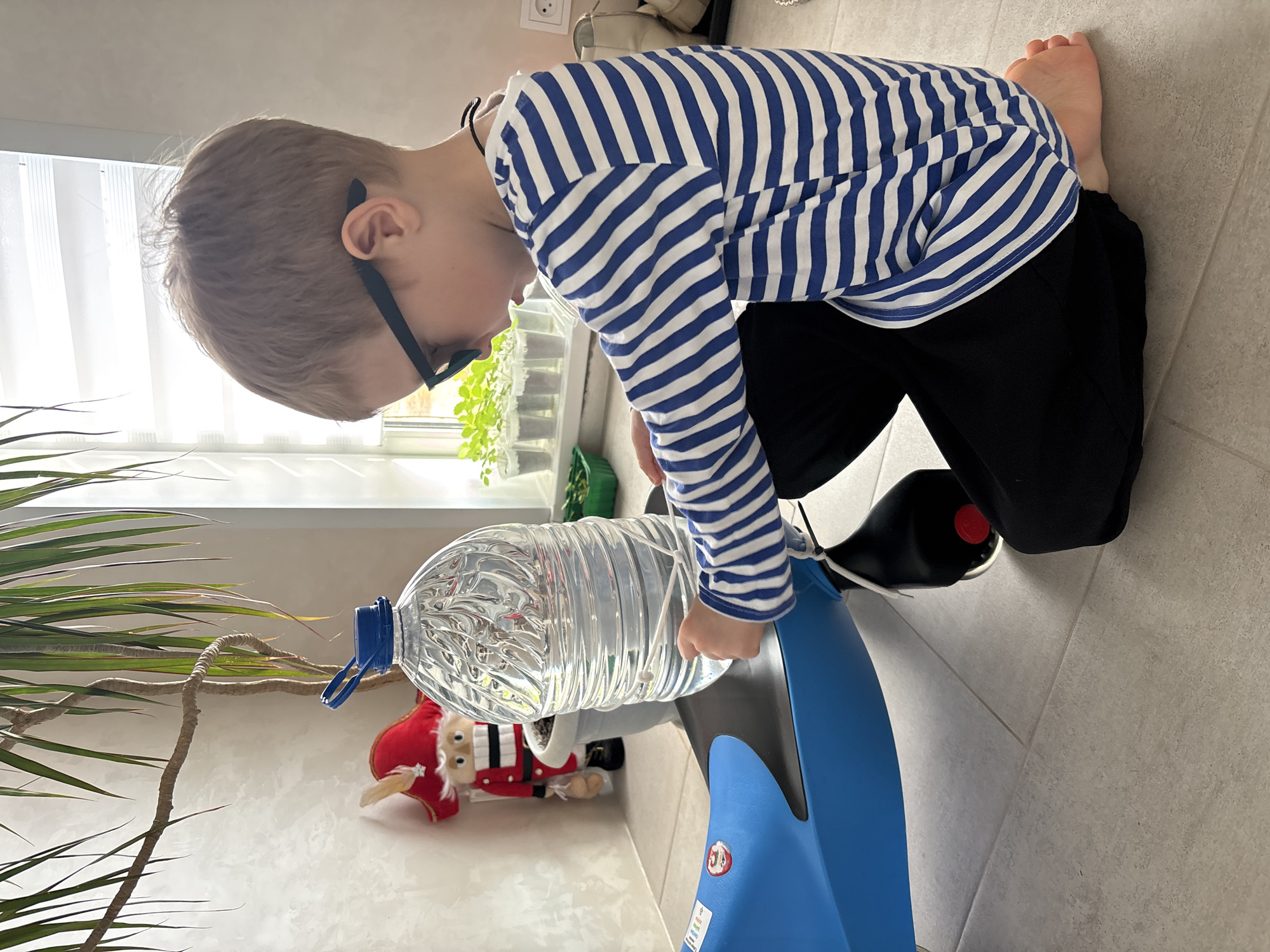 A young boy with light brown hair and black-rimmed glasses kneels on a light gray tiled floor, focused on a blue and gray water cooler. He is wearing a blue and white striped long-sleeved shirt and black pants, and is barefoot. His hands are near the top of the cooler, manipulating a large, inverted plastic water bottle that is placed on the dispensing unit.
To the left, a large green houseplant with long, narrow leaves is visible. In the background, bright daylight streams through a window covered by white blinds, illuminating a windowsill where several small potted plants or seedlings in clear containers are arranged. Below the windowsill, a decorative Santa Claus figurine in a red suit and hat, alongside another doll resembling a nutcracker, rests on a surface.
The scene depicts an indoor setting, likely a home, during daytime. The boy appears to be engaged in a task, possibly changing or adjusting the water bottle on the dispenser. On the side of the blue water cooler, a red circular sticker featuring the number "3" in white is visible. The boy's posture and gaze suggest concentration.