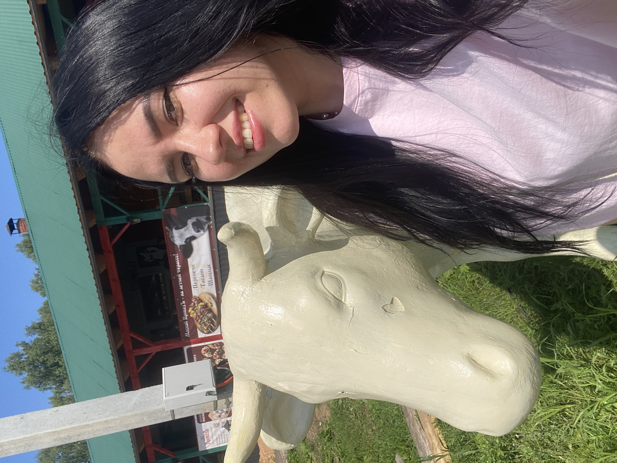 A young woman with long, dark hair and a light pink t-shirt smiles at the camera, taking a selfie. She is standing next to a cream-colored statue of a cow. In the background, there is a building with a green roof and red trim, which appears to be a restaurant or cafe, possibly with outdoor seating. Signs are visible on the building, one featuring an image of kebabs and another with text in Cyrillic. The setting is outdoors, with grass and trees visible. This location is in the city of Verkhnii Liuk, Russia.