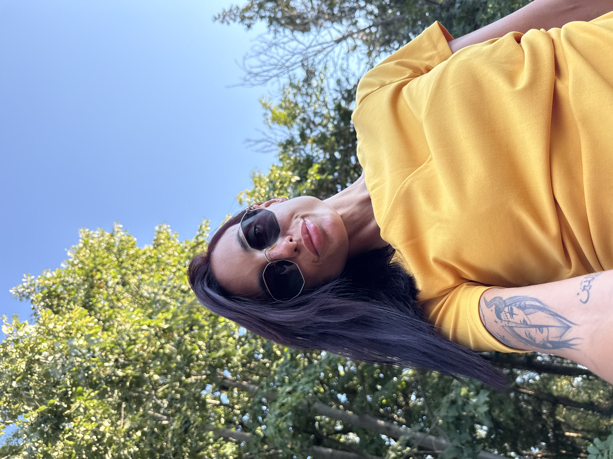 A woman with dark hair and sunglasses is shown from a low angle, outdoors. She is wearing a yellow shirt and has a tattoo on her arm. The background consists of trees and a clear blue sky. The scene is a selfie taken in a park-like setting.