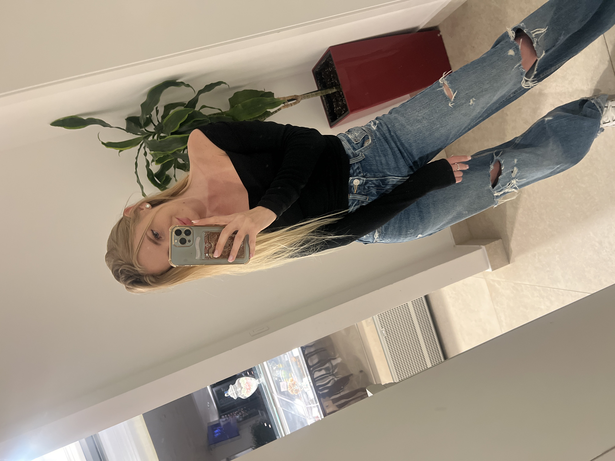 The image shows a young woman taking a selfie in an indoor environment. She is wearing a black long-sleeved top and ripped blue jeans, and she is holding a smartphone in her right hand. The woman has blonde hair.
Behind the woman is a plant in a red rectangular pot. In the background and beneath the woman, there appears to be a display case with pastries. The lighting suggests an indoor location, likely a store or restaurant. This is probably not in the city of Boreti, Montenegro, as that city is not known for its indoor shops. The floor is tiled.