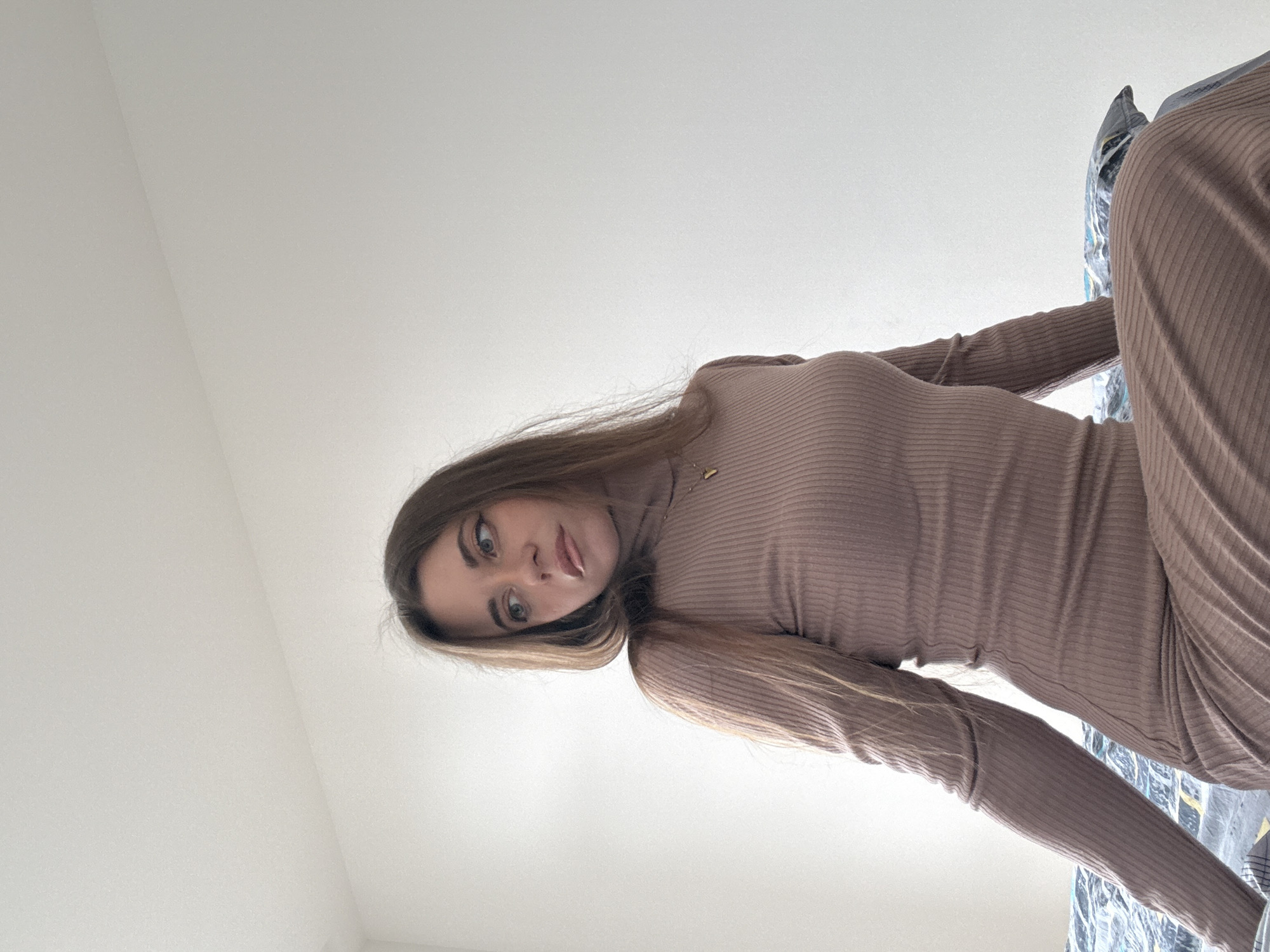 A young woman with long brown hair and green eyes, wearing a brown turtleneck dress, is looking down at the camera from an elevated position. She appears to be indoors, with a plain white ceiling visible above her. Part of a patterned bed covering is visible in the background. The image does not show any features related to the city of Moscow, Russia.