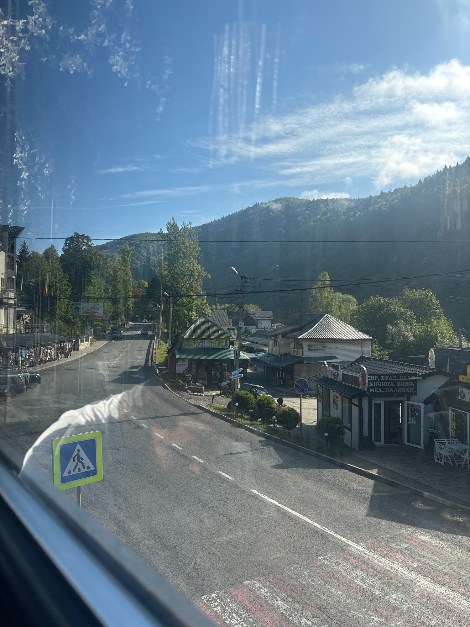 The image captures a street scene in Yaremche, Ukraine, on a clear day with a bright blue sky and scattered clouds. The perspective is from inside a vehicle, with reflections on the window obscuring parts of the view. A two-lane asphalt road runs through the center of the frame, with a crosswalk visible in the foreground. On the left side of the road, a multi-story building with apartments or hotel rooms is situated, with a market stall displaying various goods along its base. On the right side of the road, several small shops are lined up, some featuring awnings and advertising signs in Ukrainian. One sign for a shop reads "СИР, БУДЗ, САЛО, ДИЧИНА, ВИНО, МЕД, НАЛИВКИ", indicating that it sells cheese, bacon, venison, wine, honey, and tinctures. Trees and lush green hills form the backdrop, suggesting the town is nestled in a mountainous region. Several cars are visible on the road, and a few people are scattered along the sidewalk near the market. A pedestrian crossing sign is prominently displayed on the left side of the road. The overall impression is a lively street in a scenic, tourist-oriented town.