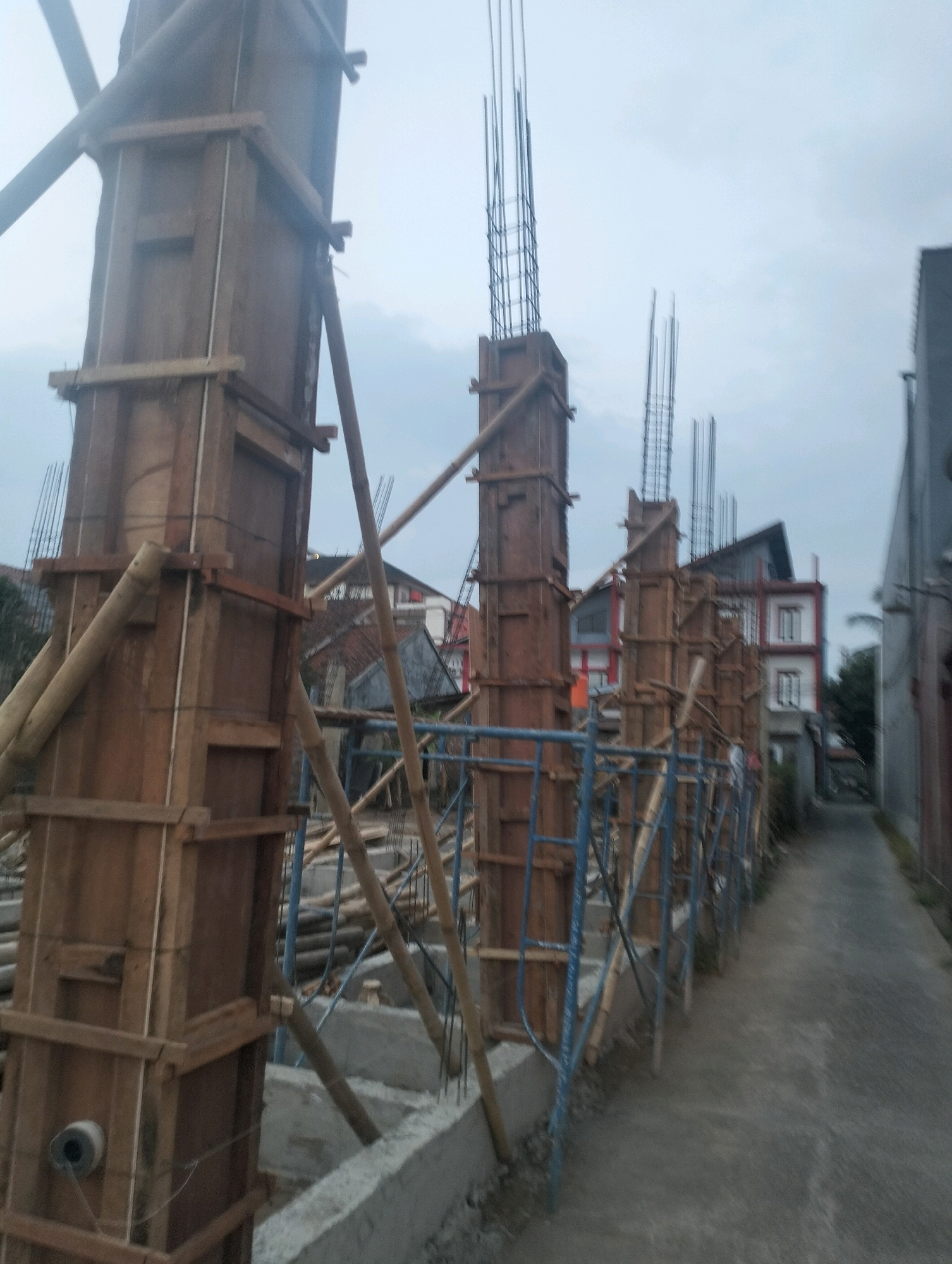 This is a photograph of a construction site in Bojongsari, Indonesia. The image shows several wooden formworks for concrete pillars, with rebar protruding from the top of some of them. The formworks are supported by bamboo poles and scaffolding. In the background, there are several residential buildings. The ground is a paved pathway. The sky is overcast, suggesting it is either early morning or late afternoon. There are no people visible in the photograph. The scene depicts the early stages of construction.