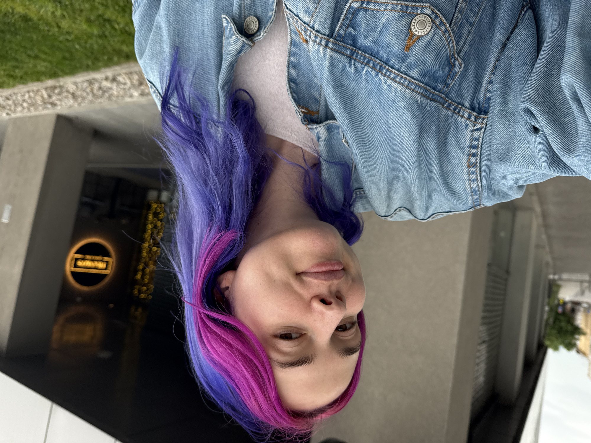 A woman with dyed purple and pink hair is standing outside in what appears to be City of Zagreb, Croatia. She is wearing a light blue denim jacket over a light colored shirt. Behind her is a building with a dark, reflective facade featuring a circular sign. The surrounding area includes concrete and greenery. The photo appears to be taken at an angle.