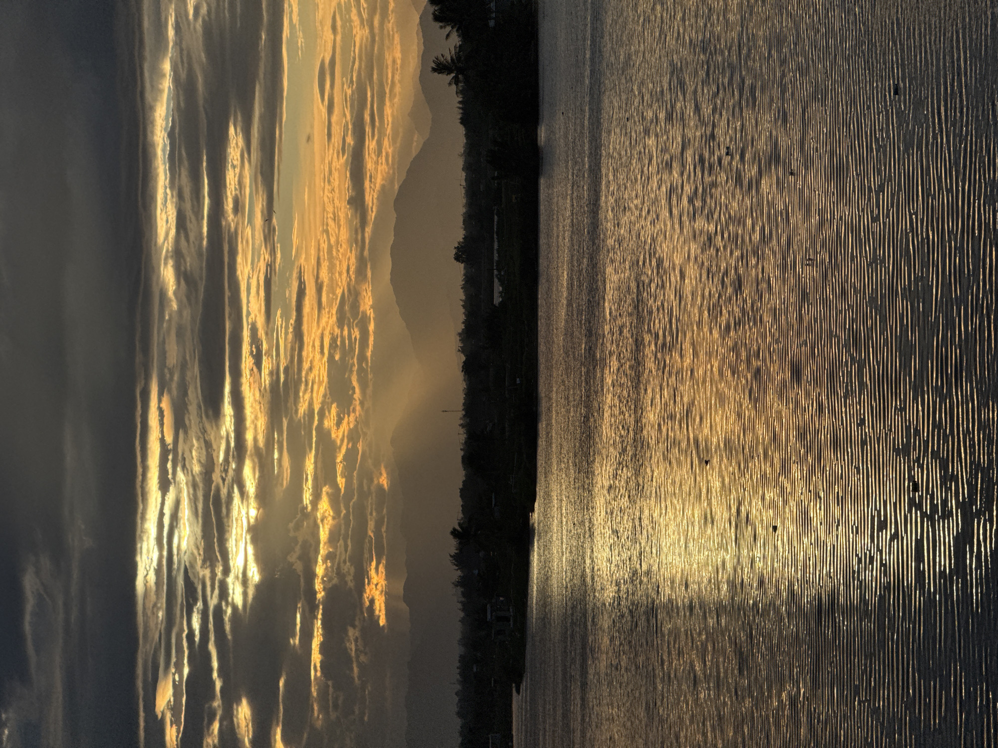 The image shows a body of water in Hoi An, Vietnam, during what appears to be sunset or sunrise. The sky is filled with dramatic clouds, with streaks of golden light breaking through, illuminating the scene with an orange and yellow glow. In the distance, silhouetted mountains form the horizon, with a dark, tree-lined shore visible in the middle ground. The water is calm, with ripples reflecting the light from the sky, creating a shimmering effect. There are no people or specific structures clearly visible in the image, making it a serene and natural landscape shot. The overall impression is one of tranquility and natural beauty.