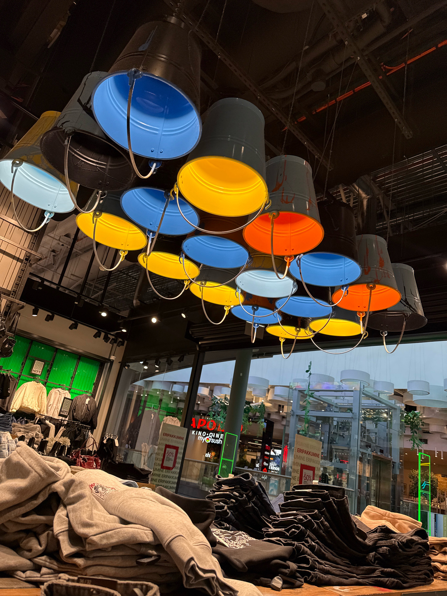 The image captures a ceiling full of colorful bucket-shaped light fixtures hanging in what appears to be a retail store. The fixtures are in various colors including blue, yellow, orange, and black. Below the lights, clothing racks with apparel are visible, along with stacks of folded clothes on display tables. In the background, there are signs with text, some of which are partially obscured. One sign reads "ERIPAKKUMINE" and another has "50%" indicating a sale. A large glass-enclosed structure, possibly an escalator or elevator shaft, is also visible. The environment suggests an indoor shopping mall or department store. The presence of retail displays and sale signs points to a commercial establishment. Based on the "ERIPAKKUMINE" and the general aesthetic, the location is likely a retail store in Tallinn, Estonia. There are no visible people in the image.