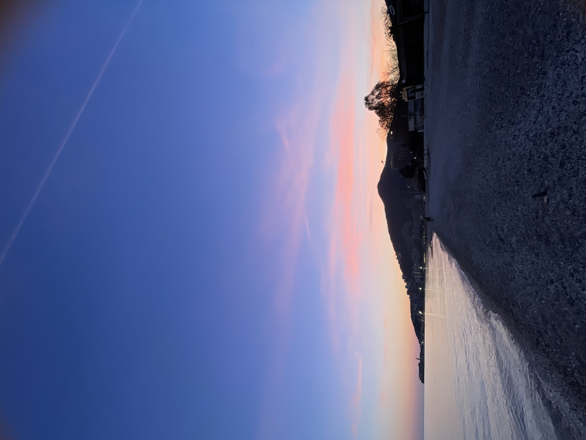 The image depicts a coastal scene in Budva, Montenegro, observed during twilight. The upper sky transitions from deep blue overhead to a vibrant blend of light blue, lavender, pink, and orange along the horizon. A single, faint contrail extends diagonally across the upper right quadrant of the sky. The Adriatic Sea occupies the left mid-ground, presenting a calm, dark grey-purple surface with minimal wave activity breaking onto the shore.
The foreground is dominated by a wide, dark grey pebble beach extending from the bottom of the frame towards the distant coastline. Two indistinct human figures, visible only in silhouette, are present on the pebble beach in the mid-ground, walking towards the right side of the frame.
The distant coastline to the left features scattered city lights and a prominent, illuminated structure resembling a lighthouse or a distinct building. The background to the right is anchored by a large, dark mountainous landmass, its lower slopes containing numerous buildings and scattered artificial lights. Silhouetted trees and low-rise structures line the immediate edge of the beach on the right side of the image.
