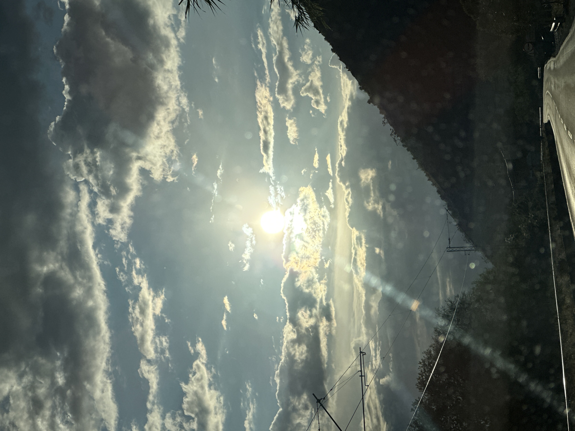 The image captures an expansive sky and a natural landscape in Vranjina, Montenegro, likely seen through a vehicle window. The sky dominates the upper and left portions of the frame, filled with a dynamic arrangement of clouds. The sun, a brilliant disc, is positioned in the upper-left quadrant, partially veiled by clouds. Its intense light illuminates the edges of the clouds with golden hues and produces prominent lens flares and sunrays that stretch across the sky. Some clouds are dense and dark, contrasting with the brighter, elongated formations that glow from the sun's light.
Towards the right side of the image, a dark, tree-covered hillside or mountain rises, bordering a paved road with a clear white line visible at the bottom-right. In the lower-left to mid-left, utility poles and electrical wires crisscross the frame. Dark foliage, possibly from a tree close to the camera, is visible along the very top-left edge. Numerous circular and streak-like reflections and small specks, indicative of dust or water on a window, are present throughout the image. The dramatic lighting, with the sun low in the sky, strongly suggests either sunrise or sunset, creating a powerful and atmospheric scene. No people or visible text are present.