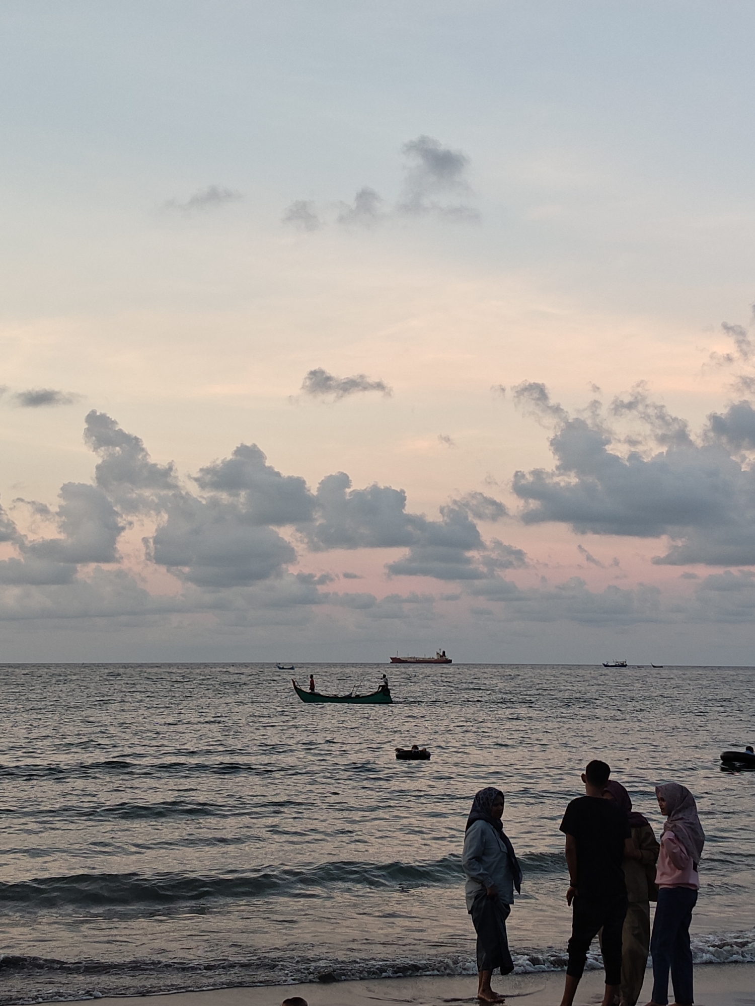The image depicts a beach scene in Lhokseumawe, Indonesia, at dusk. A group of four people stands near the shoreline; two women wear hijabs. In the background, the ocean is visible with several small boats and a larger ship. The sky is filled with clouds tinted with shades of pink and gray.