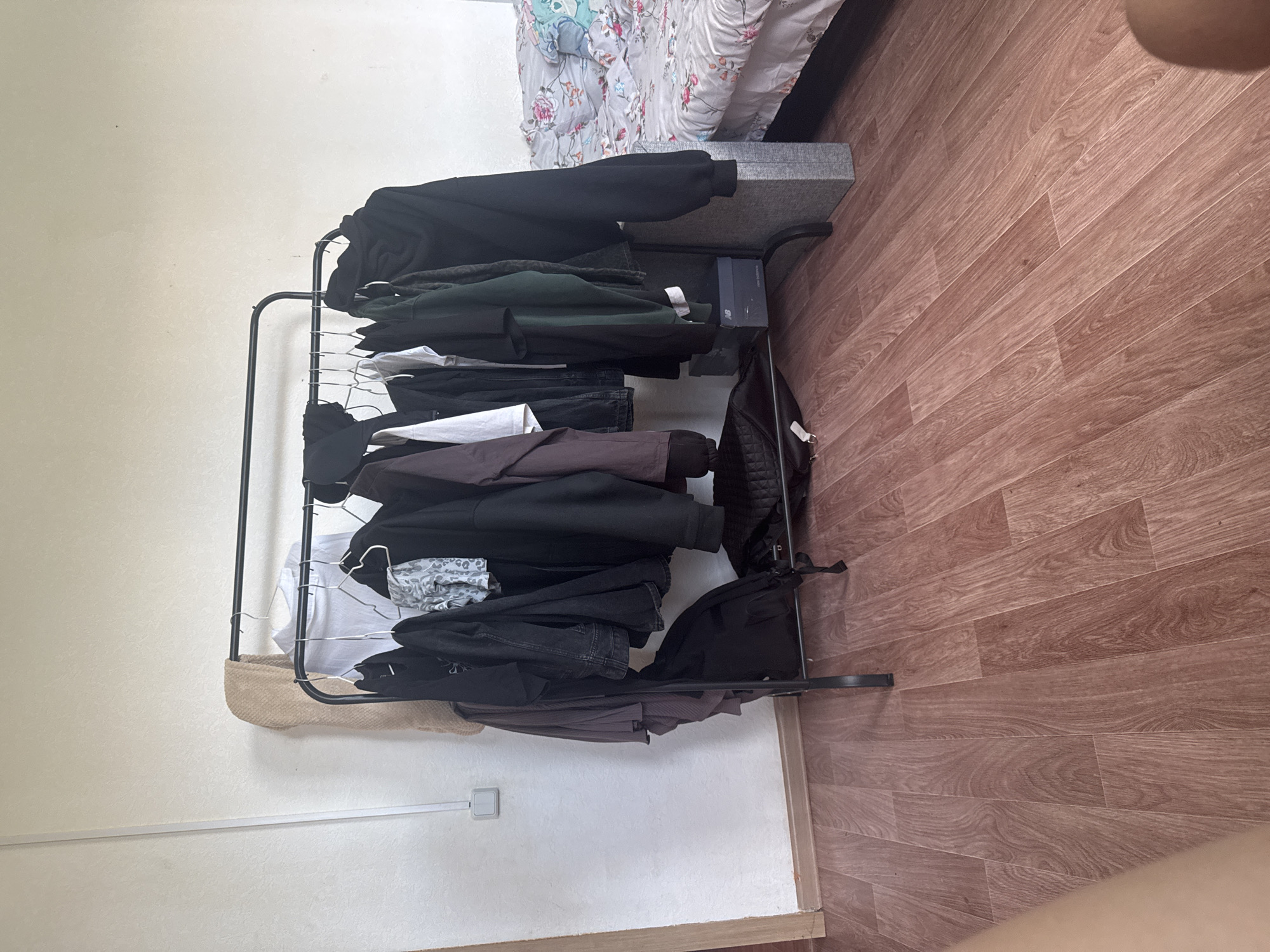 The image shows a bedroom in an apartment. On the left, there is a white wall with a light switch. In the center, there is a black metal garment rack with various clothes hanging on it, including jackets, shirts, and a blanket. Below the rack, there are bags and boxes. To the right, there is a bed with a floral patterned comforter and pillows. The floor is covered with wooden laminate flooring. The room appears to be located in the city of Krasnyye Baki, Russia.