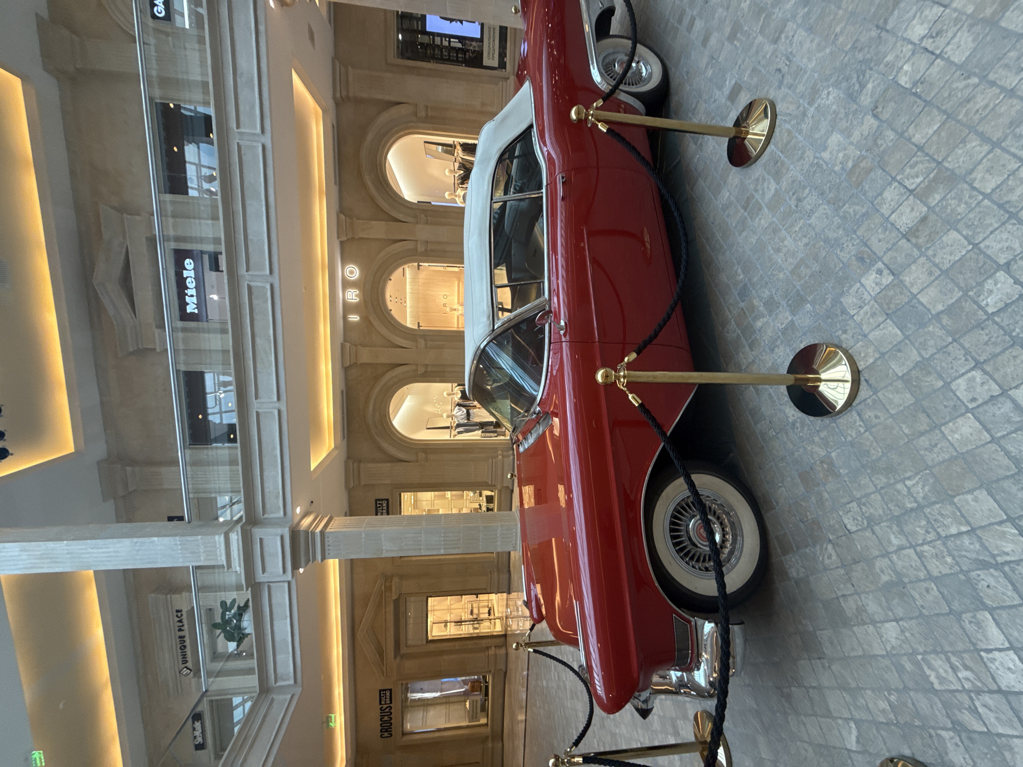 The image shows a vintage red convertible car with a white roof parked on a stone-tiled floor. The car is positioned diagonally, with its front end slightly obscured. It is roped off with stanchions and black rope, suggesting it's an exhibit or display.
The background reveals an indoor setting that appears to be a high-end shopping mall or a large public building. Architectural elements like arched doorways, columns, and multi-level facades are visible. Stores with illuminated signage are integrated into the architecture. Text visible in the background includes "Miele", "UNIQUE PLACE", "IRO", "MULTI BRAND", and "CROCS". The lighting suggests it is daytime, with warm light illuminating the interior architecture. There are no people visible in the immediate vicinity of the car, contributing to a sense of stillness and focus on the vehicle. The overall impression is one of elegance and a display of automotive history within a sophisticated environment.