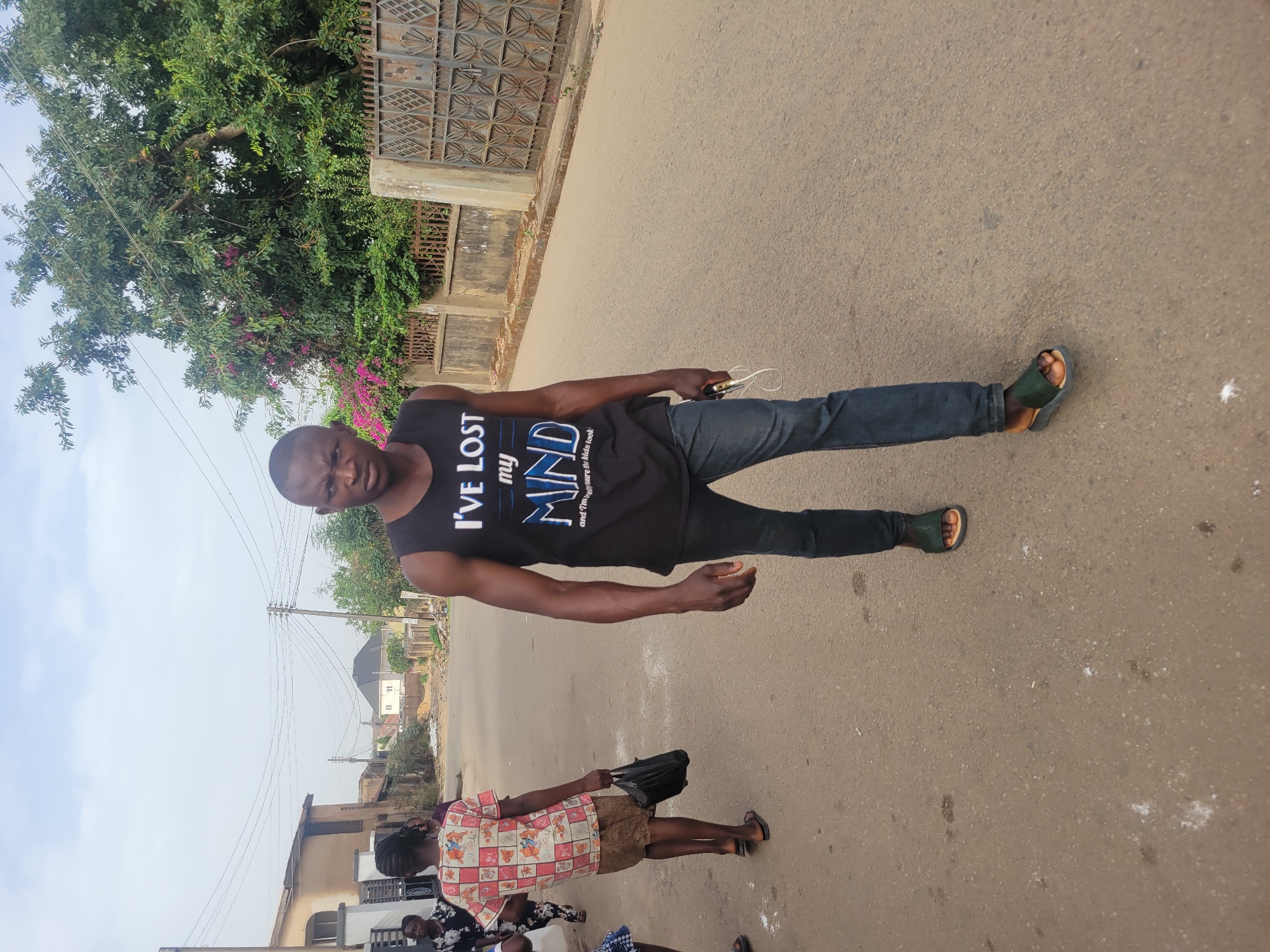 A medium shot captures a man in Jos, Nigeria, standing on a paved road. He wears a black tank top with white and blue text, dark jeans, and dark green sandals. He holds a white wired earphone in his left hand. To his right, along the roadside, is a fenced property with trees and pink flowers. In the background, utility poles and buildings are visible. In the lower part of the frame, other people are partially visible.