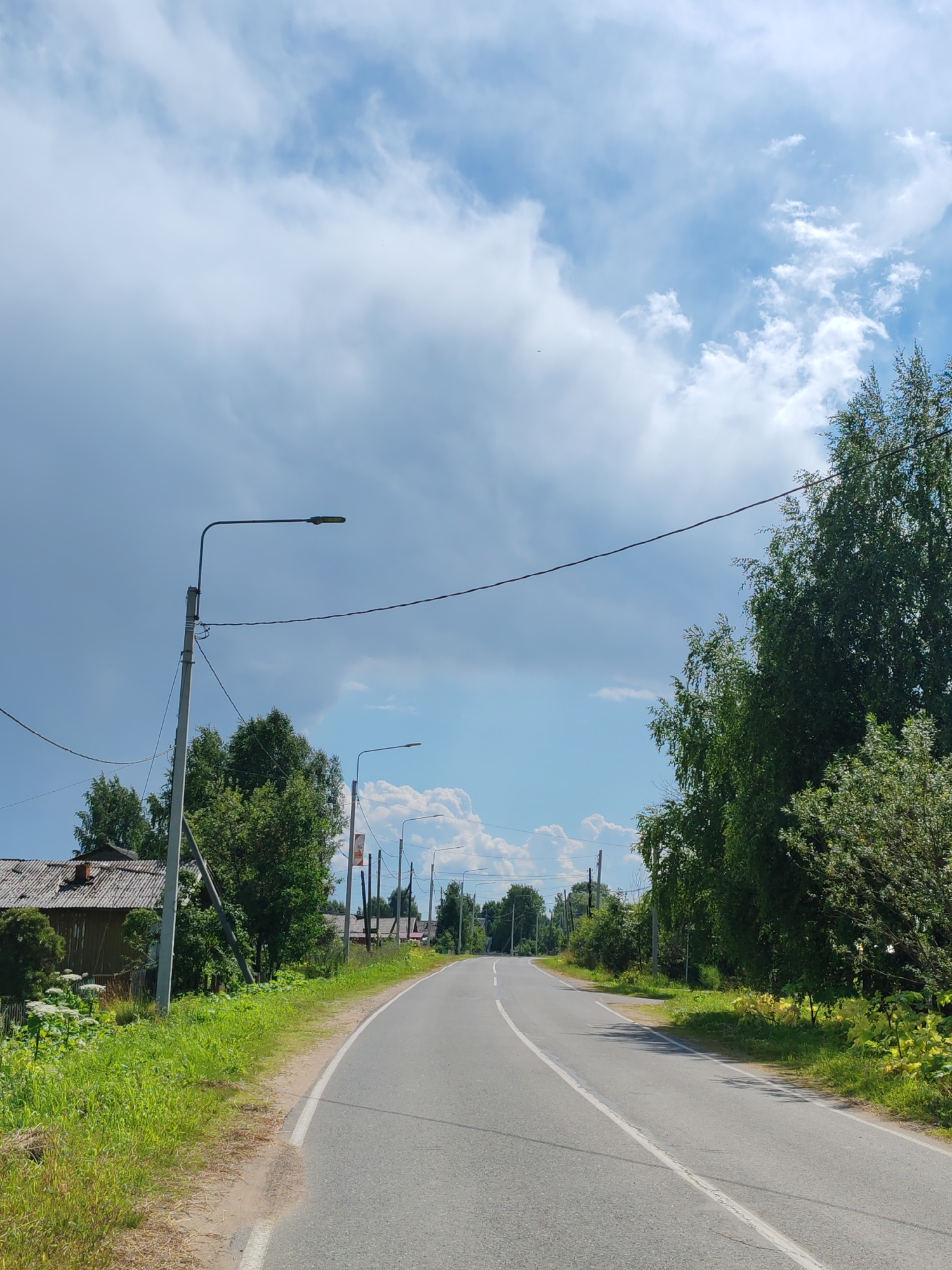 This is a vertical, eye-level, medium shot of a paved road curving to the left in the village of Chukhlem, Russia. The sky is a bright blue with scattered white clouds, some of which appear heavy and dark. Tall green trees line the right side of the road, with smaller trees and a wooden building visible on the left. A power line runs horizontally across the frame. Two streetlights are visible on poles along the road, and there are other utility poles and wires in the background. The road has a white line dividing its lanes and a white line marking the edge of the shoulder. The shoulder is unpaved and covered in grass and some yellowing vegetation. The scene is illuminated by natural daylight.