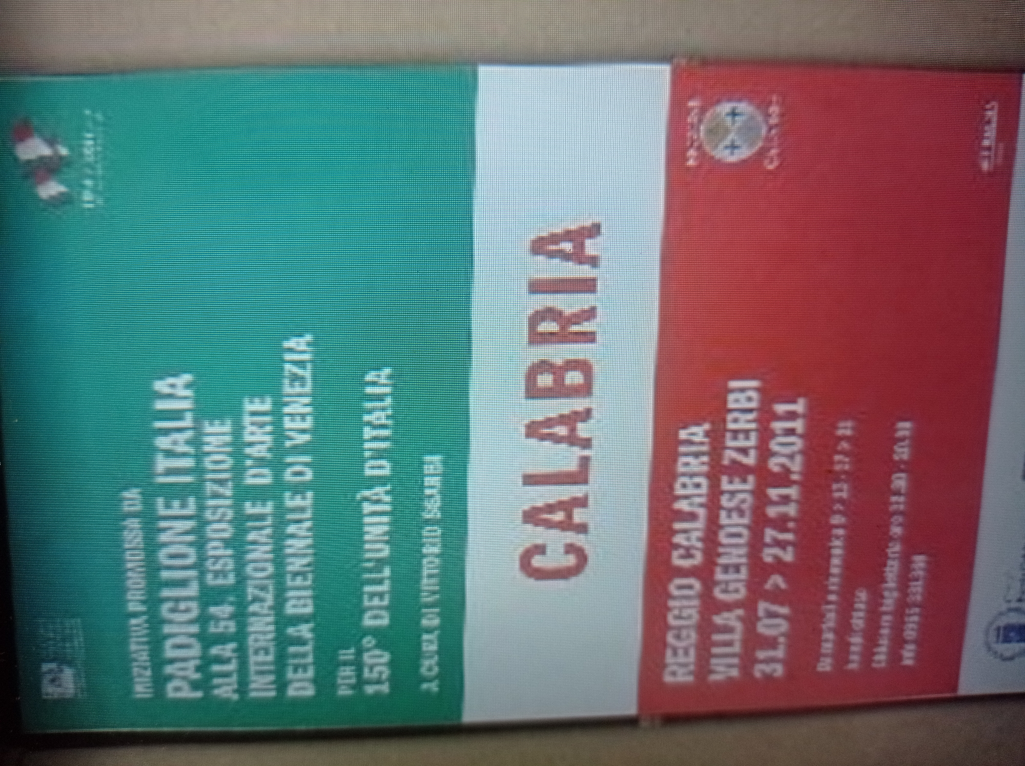 The image is a photograph of a poster or banner with Italian text. The text is displayed in white and red against a green and red background. The poster advertises an exhibition in Venice, Italy, related to the 150th anniversary of Italian unification. The lower portion of the poster indicates an event in Reggio Calabria, Italy, from July 31 to November 27, 2011, at Villa Genoese Zerbi. The image does not contain any information or visual cues related to the city of Mukachevo, Ukraine.