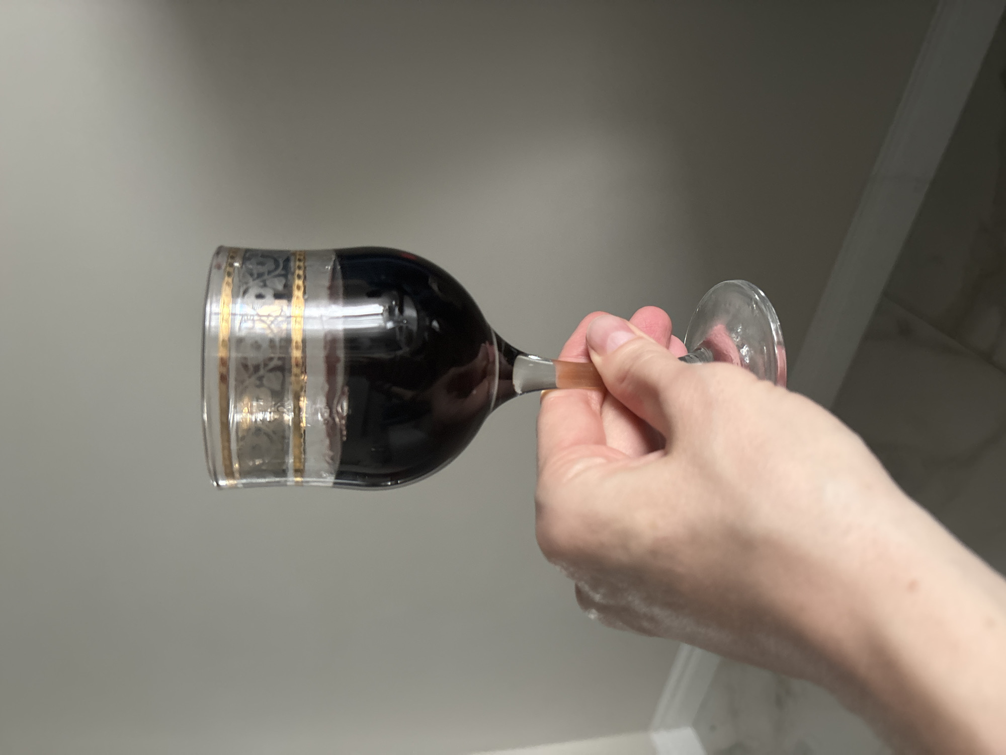 A hand holds a wine glass filled with a dark liquid against a light grey background. The glass has a decorative gold band near the top. The location is Unknown, Unknown.