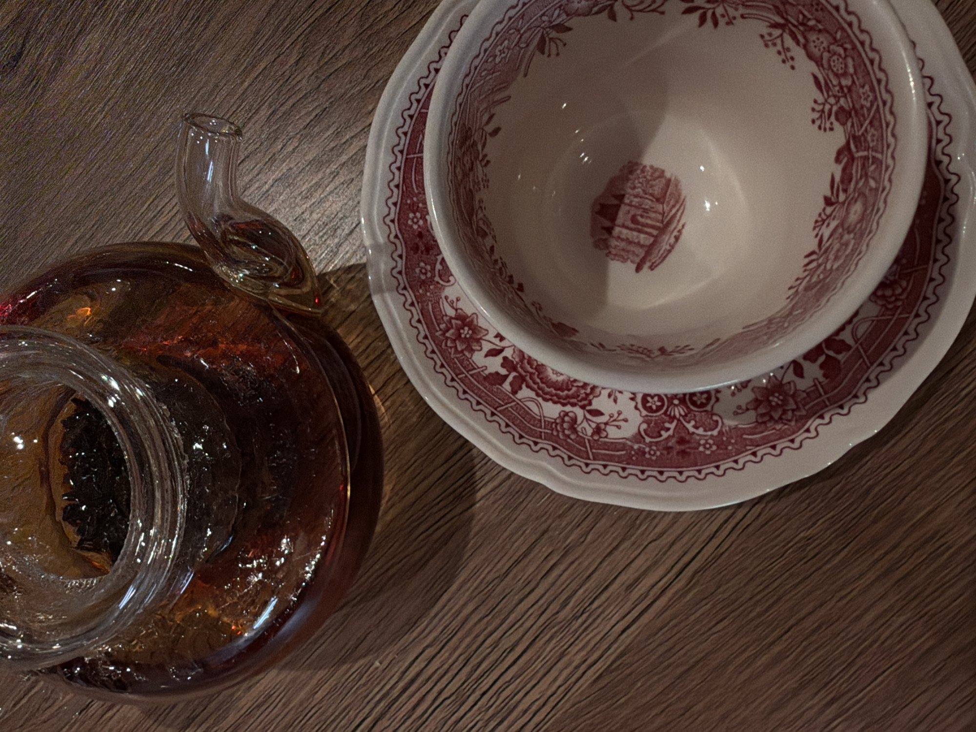 The image shows a close-up, high-angle shot of a wooden surface. Two objects are prominently featured: a transparent, amber-colored glass pitcher or jar with a curved spout, and a teacup with a saucer decorated with a red floral pattern. The objects are positioned side-by-side on the wooden surface. The teacup is placed on top of the saucer. There is no information to confirm a location in the city of Khimki, Russia.