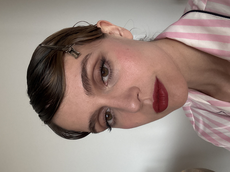 A female individual is depicted in a close-up, chest-up portrait, situated indoors within the city of Unknown, Unknown. The subject exhibits fair skin, brown eyes with visible dark mascara or false eyelashes, and dark, well-defined eyebrows. Her facial cosmetics include neutral-toned eyeshadow, distinct pink blush on the cheeks, and dark red matte lipstick on full lips. Her dark brown hair is styled with a wet, slicked-back appearance, forming a distinct wave over the forehead. A silver-toned metal duckbill hair clip is secured in the hair above her right ear. She is wearing a collared garment, likely a pajama top, featuring vertical pink and white stripes with dark piping along the collar edge. The immediate background is an unadorned, light-colored wall.