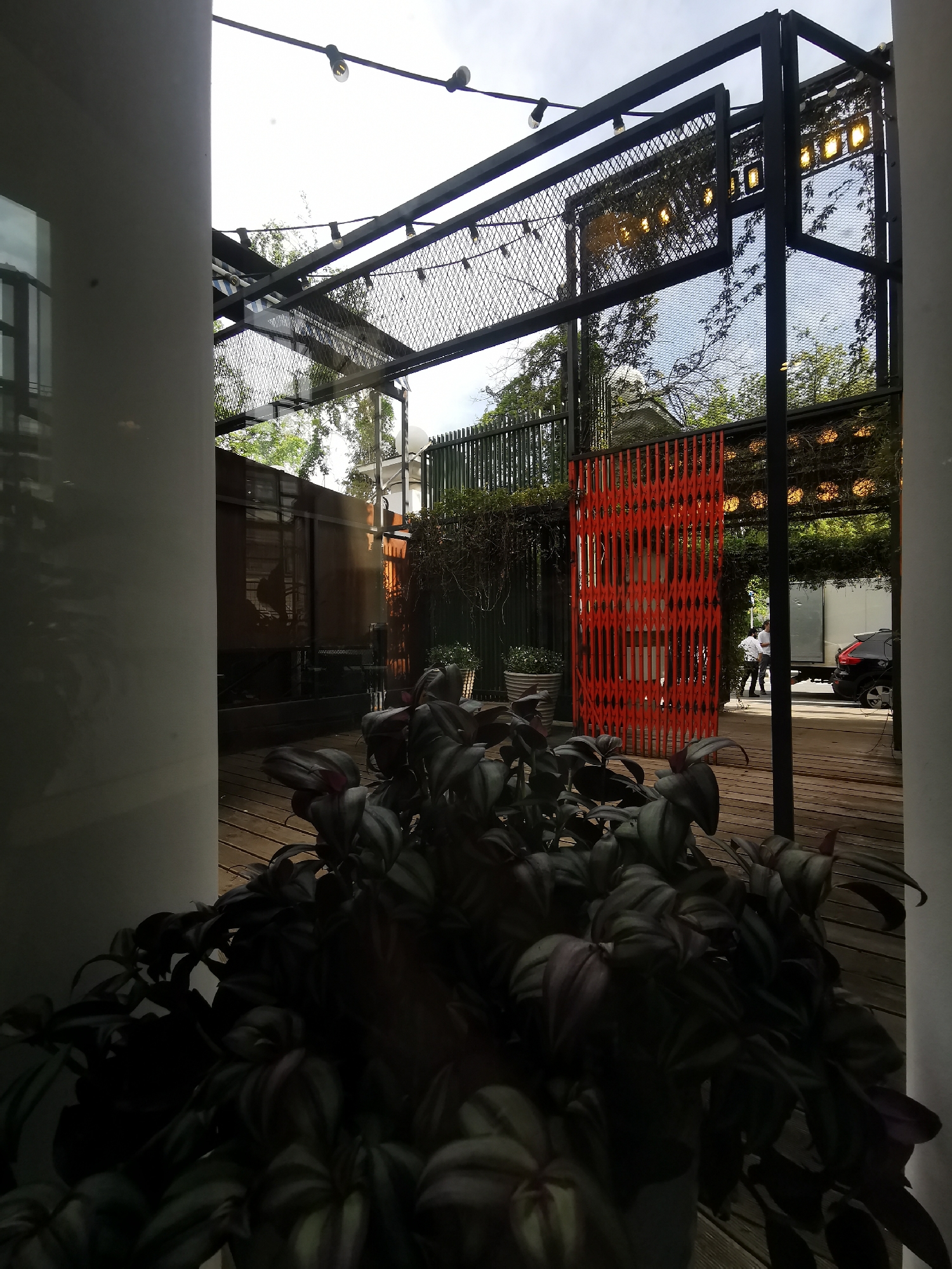A view through a building opening shows an outdoor space, with a potted plant in the foreground. The space beyond features a wooden deck, a metal pergola structure, and a retractable orange gate. In the background, two people stand near a delivery truck and a car. The exact location in Moscow, Russia cannot be determined without additional information.