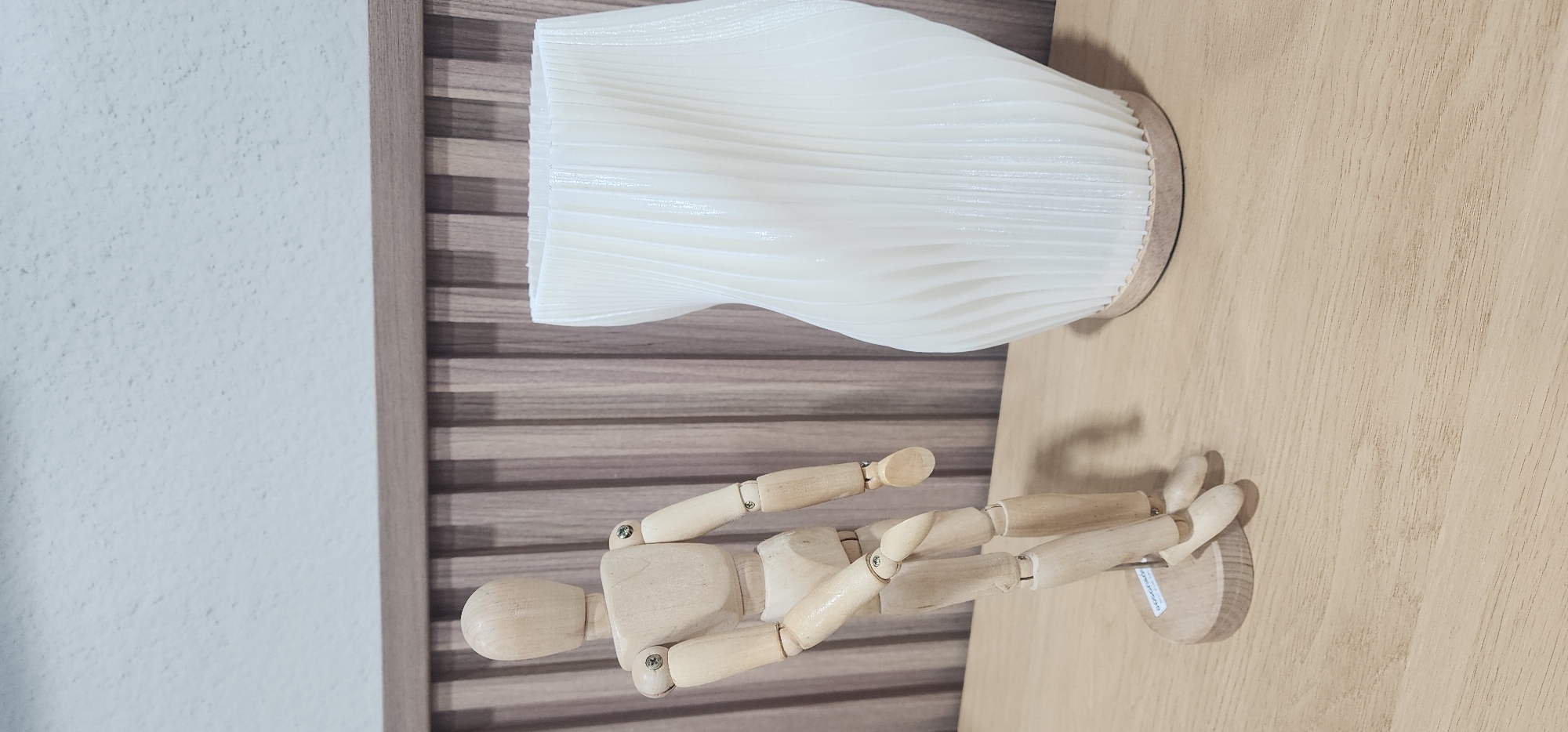A wooden articulated mannequin stands to the left of a white, twisted, cylindrical vase with a wooden base. The objects are placed on a light brown wooden surface. In the background, a wall with a textured, light blue surface is visible above a panel of vertical wooden slats. The scene is indoors, with no indication of being in Baan Chaweng Noi, Thailand or any specific city.