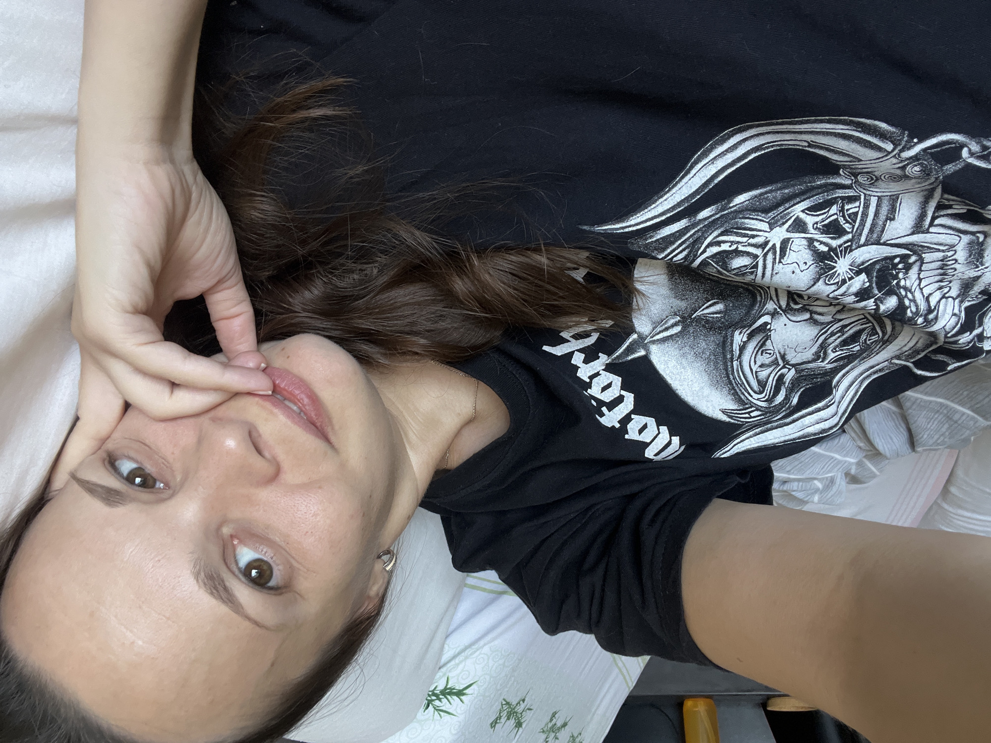A woman with brown hair and brown eyes is lying on her side in bed, looking at the camera. She is wearing a black t-shirt with a white graphic design. Her right hand is resting on her cheek, and her fingers are touching her lips. The background is a white pillow and a dark shelf with an orange bottle on it. The scene is indoors, likely a bedroom, and is not specifically identifiable as being in the city of Tolyatti, Russia.