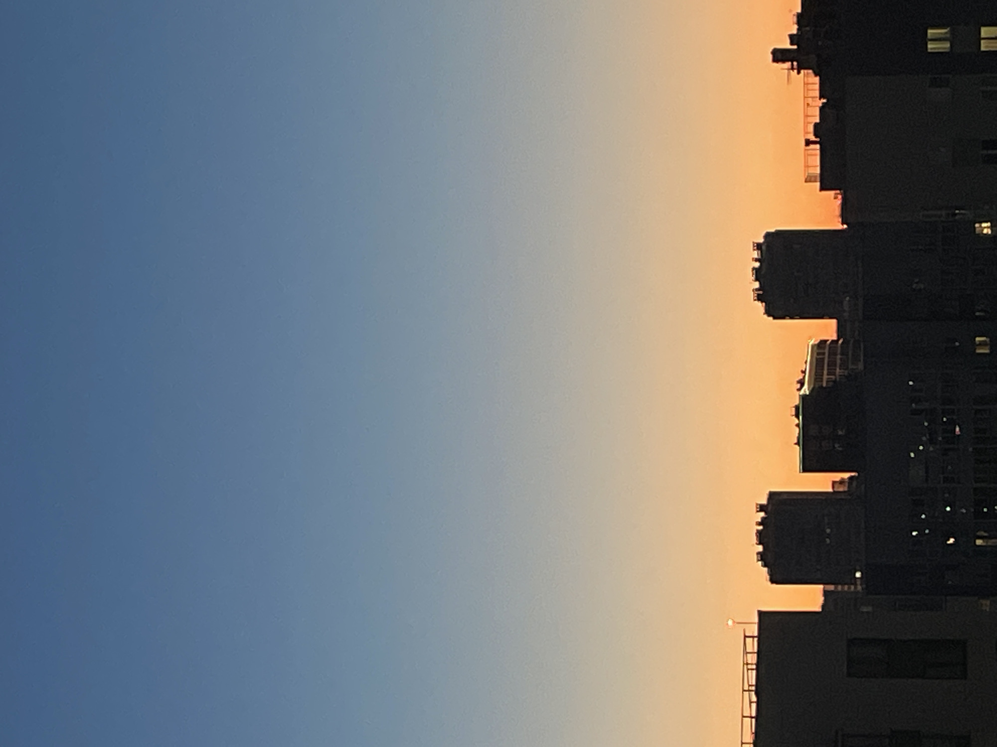 This is a vertical, medium-shot photograph of the Sinyavino, Russia skyline at dusk. The sky transitions from a deep blue at the top to a vibrant orange near the horizon. Silhouetted against the sky are several multi-story buildings, appearing as dark geometric shapes. Some buildings have visible windows or architectural details. A singular small, bright light, possibly a streetlamp, is visible on the rooftop of a building on the far left. The overall scene is one of urban stillness during twilight.