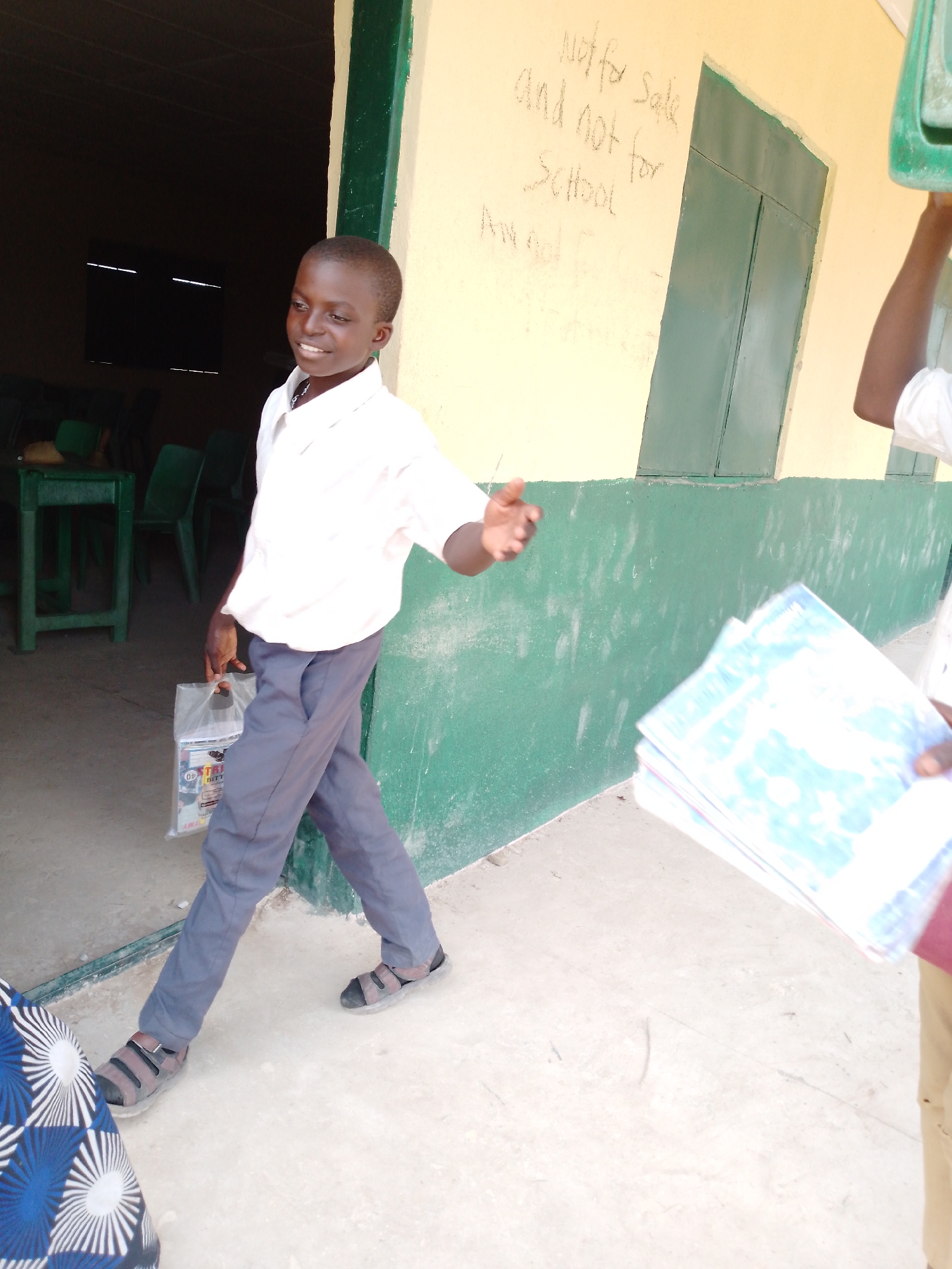 Akwanga, Nigeria. An adolescent boy in a white shirt and grey trousers walks out of a building with green-trimmed windows. He carries a plastic bag. Another person to the right holds a stack of papers or books. The building wall is painted yellow and green, with the words "Not for sale and not for School" written on it.