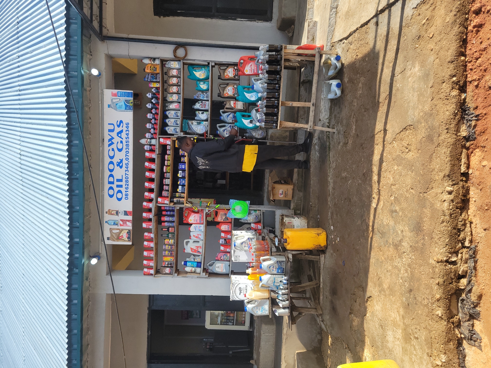 This is an outdoor, eye-level, medium shot of a small shop in Jos, Nigeria, specializing in oil and gas products. A man wearing a black jacket with a yellow shirt underneath is standing inside the shop and appears to be attending to the products. The shop is filled with shelves stocked with various containers of oil, lubricants, and other automotive fluids. In front of the shop, there are wooden tables displaying more products, including large yellow plastic containers. The ground in front of the shop is unpaved and appears dusty. The shop has a corrugated metal roof and a green trim. The overall scene depicts a busy retail environment for automotive supplies in an urban setting.