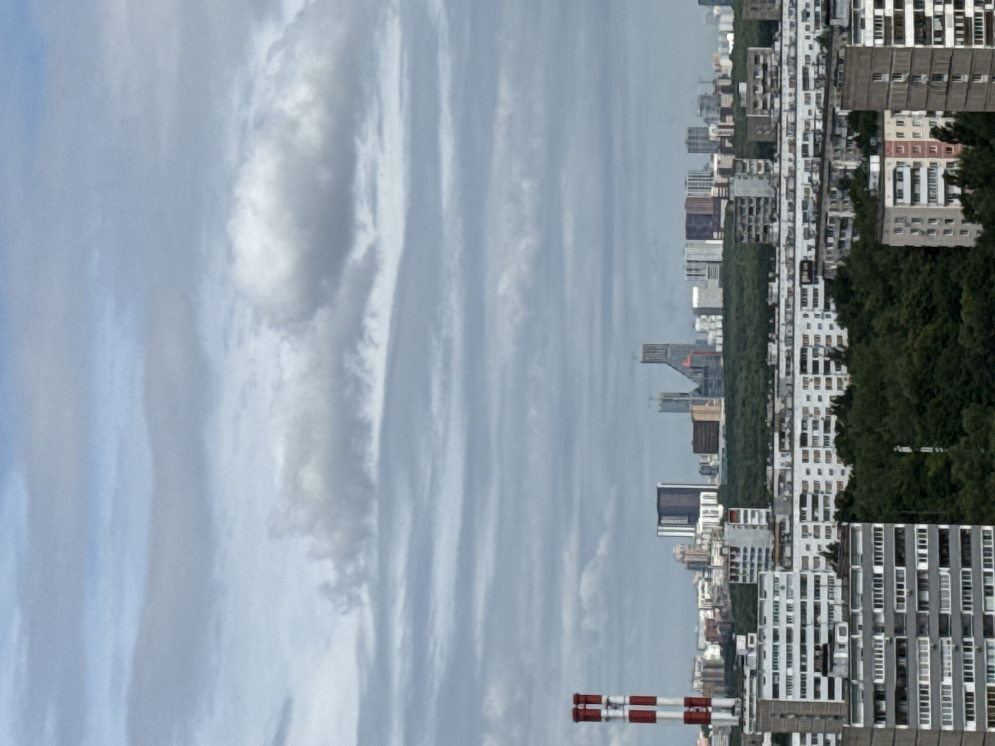 This is a high-angle, medium shot of a residential area in Moscow, Russia. The sky is overcast with a layer of gray clouds. In the foreground, there are several multi-story apartment buildings with a mix of white, gray, and beige facades. Some buildings feature balconies and numerous windows. To the left, a tall industrial chimney with red and white stripes rises into the sky. Behind the apartment buildings, a dense green forest stretches across the middle ground. In the background, a cityscape of various modern and older high-rise buildings is visible under the cloudy sky. The scene depicts a typical urban residential landscape with a mix of Soviet-era and more contemporary architecture, with a prominent industrial element.
