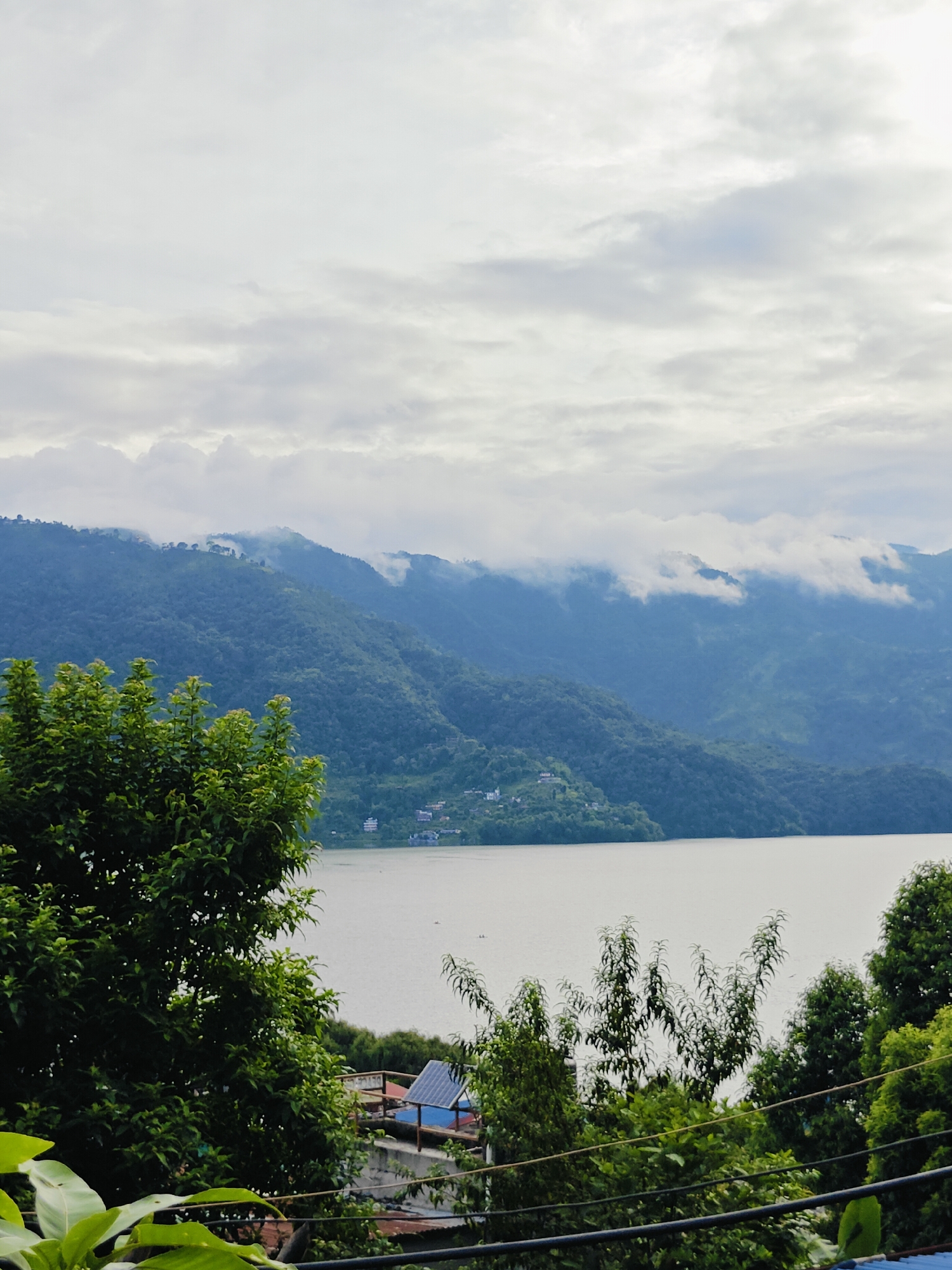 The image shows a scenic view of Phewa Lake in Pokhara, Nepal, with lush green hills and a cloudy sky. In the foreground, there are dense trees and foliage. Below the trees, a few buildings with red roofs are visible, along with a solar panel on one of the rooftops. The lake itself appears calm and reflects the overcast sky. The hills in the background are covered in vegetation, and clouds are seen partially obscuring the peaks, creating a dramatic and misty atmosphere. There are no people visible in the scene.