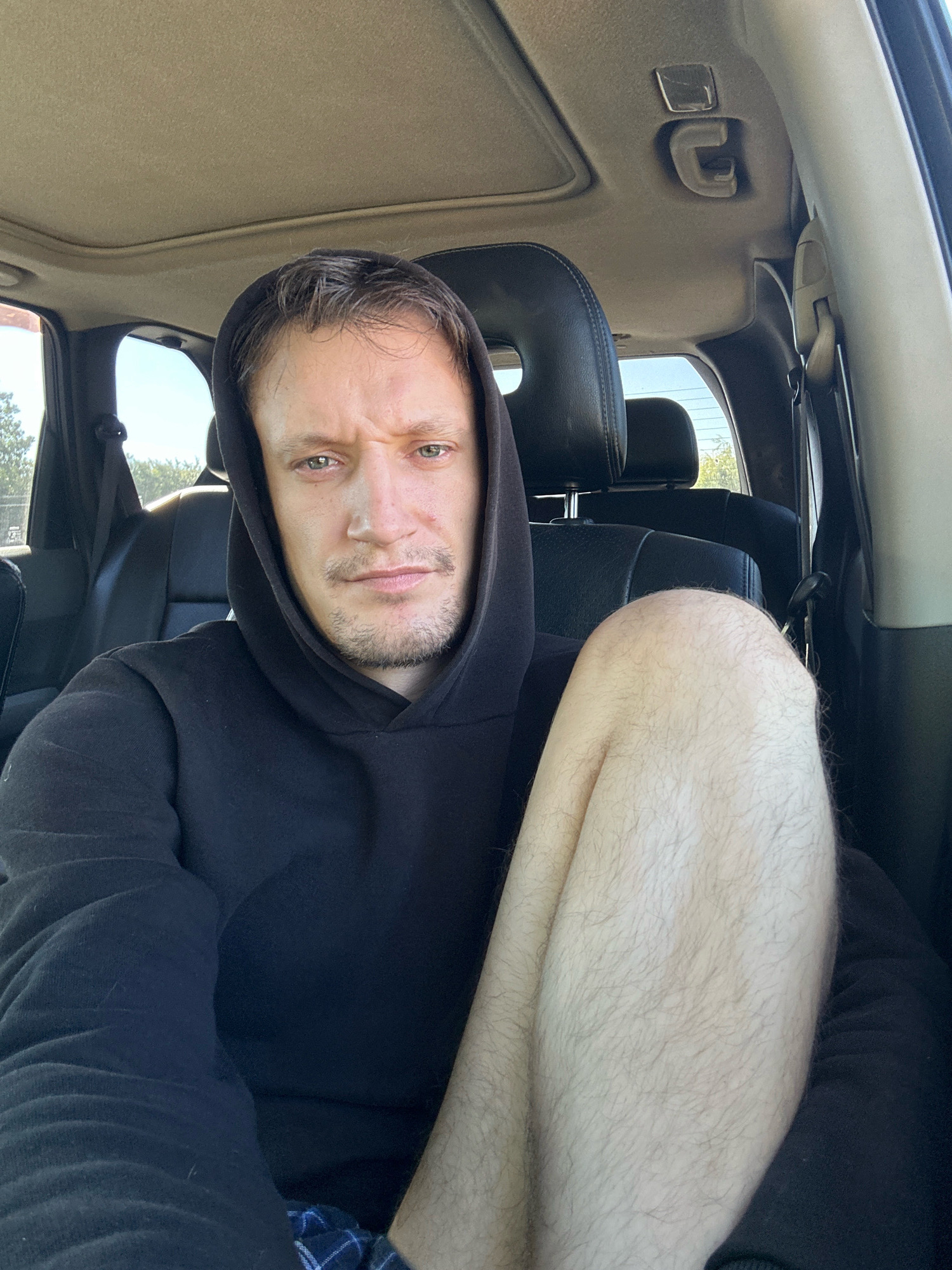 A man wearing a black hoodie is sitting in the passenger seat of a car. He has light skin, green eyes, and short brown hair. His left leg is crossed over his right leg. The interior of the car is visible, including black leather seats, the sunroof, and the roof lining. The car appears to be parked, possibly in the city of Makiv, Ukraine, as suggested by the natural lighting and foliage visible through the windows. The man is looking directly at the camera with a neutral expression.