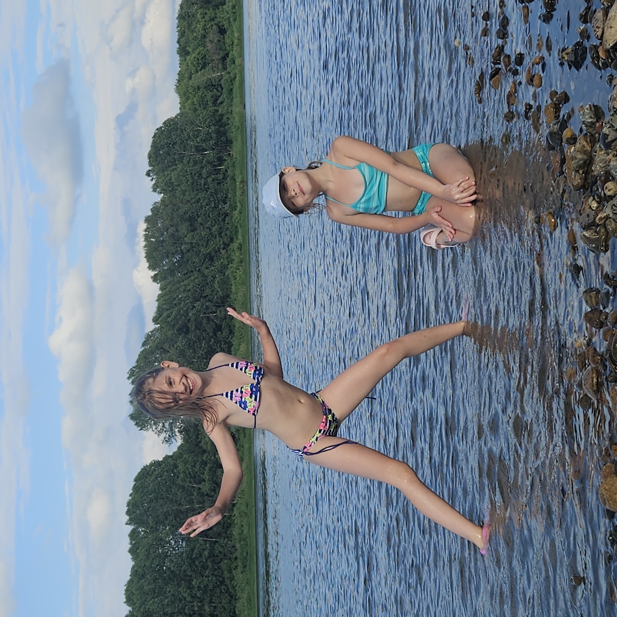 Two young girls are in the water near a rocky shore, with trees and a cloudy sky in the background. The girl in the foreground is wearing a colorful bikini and has her arms outstretched, appearing to be in motion. She has a wide smile and her hair is wet. The second girl is further back in the water, wearing a turquoise bikini and a white cap. She is sitting and looking towards the camera with a calm expression. The water is clear and rippling, and the sun appears to be shining. The scene suggests a playful moment during a summer day at a lake or river in Elykaevo, Russia. There is no visible text in the image.
