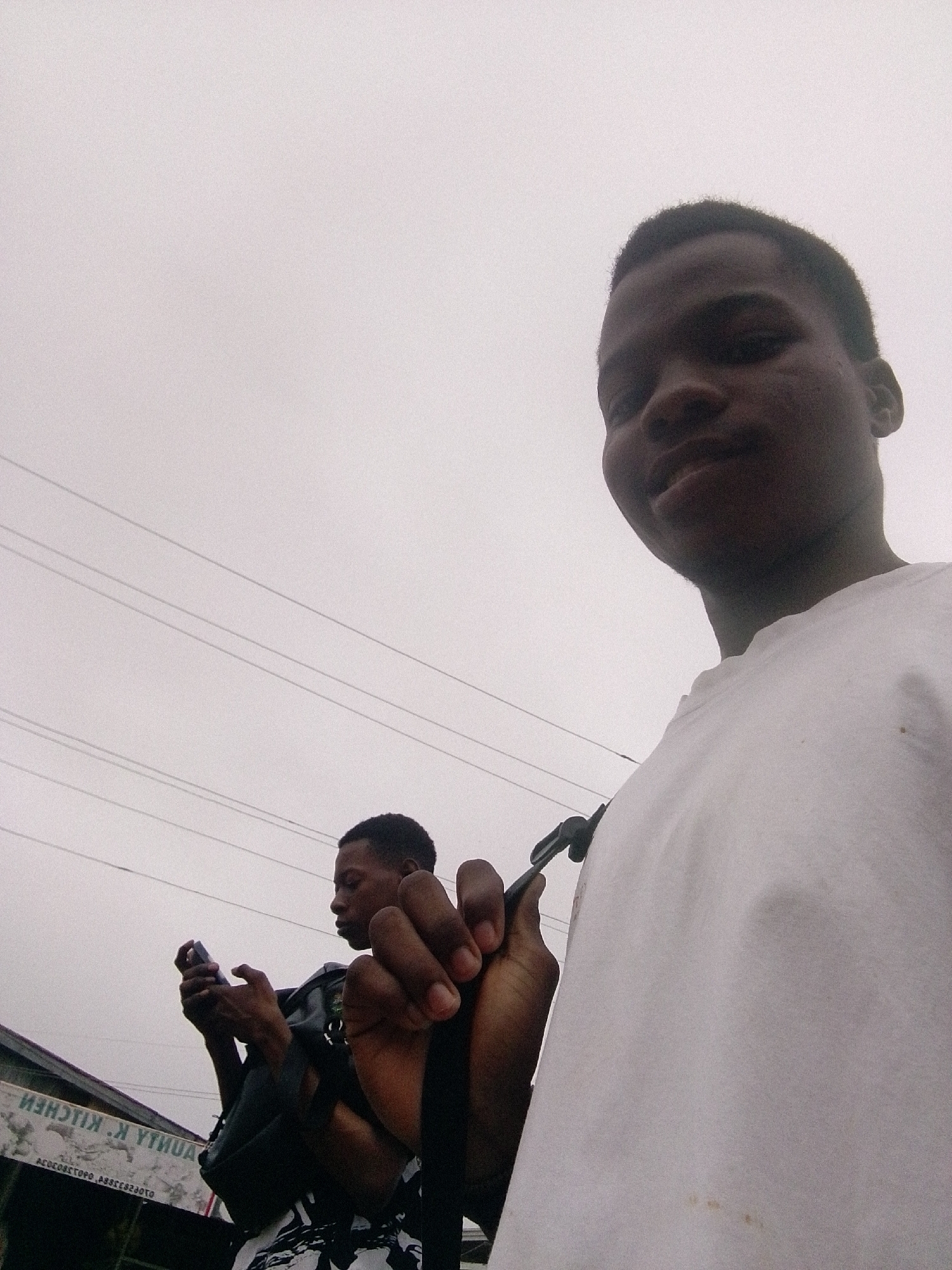 Two young men are visible in the image. The main subject is closer to the camera, wearing a white shirt and holding a black object. His features are clearly visible. Behind him, another young man is seen using a mobile phone. He is wearing a dark shirt and a backpack. The background suggests an urban environment, featuring overhead power lines and a partial view of a building with signage that reads "IYA KITCHEN" in reversed lettering, indicating the location is likely near or adjacent to a food establishment in Ipetumodu, Nigeria. The sky is overcast.