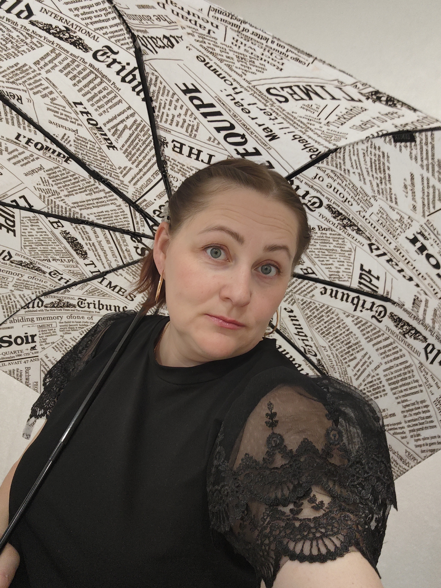 A woman with fair skin, blue eyes, and auburn hair pulled back is holding a black and white newspaper print umbrella indoors. She wears a black top with lace sleeves and gold hoop earrings. The background is plain and white. The text on the umbrella includes headlines like "INTERNATIONAL," "Tribune," "TIMES," and "L'EQUIPE" in a variety of fonts. The specific location is not discernible from the image.