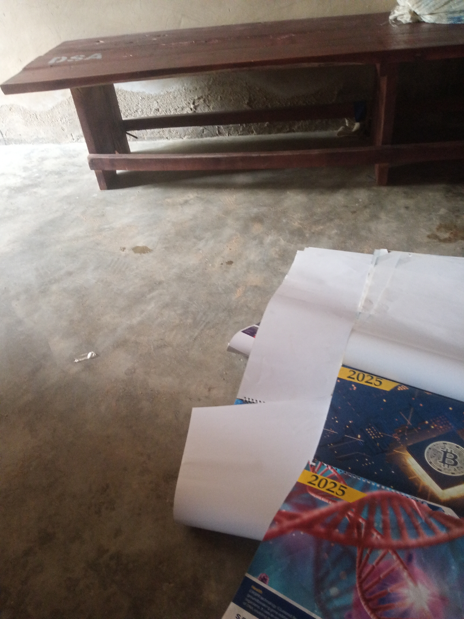 The image shows a wooden desk with the letters "DSA" stenciled on its surface. The desk appears to be placed on a concrete floor, possibly in an indoor or covered outdoor setting. The floor has some marks and stains, suggesting it is an everyday use area. In the foreground, several sheets of white paper are stacked or rolled, partially obscuring some items below. The visible items beneath the paper appear to be a magazine or book with the year "2025" prominently displayed on its cover in a yellow band. The cover also features imagery of a DNA helix and what looks like a Bitcoin logo. The overall scene suggests a casual or academic environment where study materials or publications are kept. The lighting indicates it could be daytime, though the specific time of day is not discernible. No people are visible in the image. The setting does not provide strong cues for weather conditions. The location context suggests this scene is in Jalingo, Nigeria.
