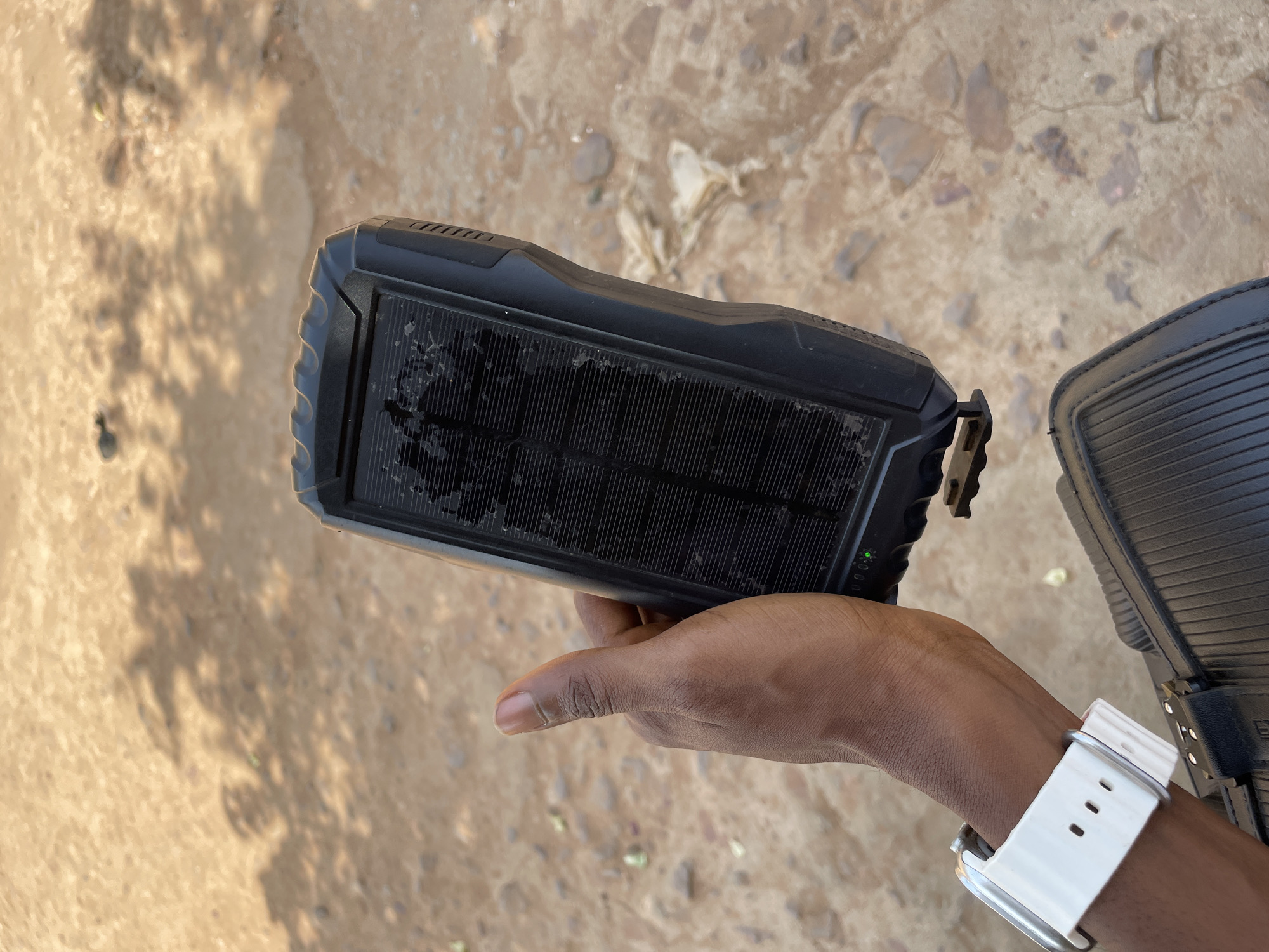 A hand is holding a small solar panel in Obosi, Nigeria. The panel is rectangular with a black frame and a ridged surface. The hand holding the panel is dark-skinned and has a white watch on the wrist. The background is a rough, light brown surface.