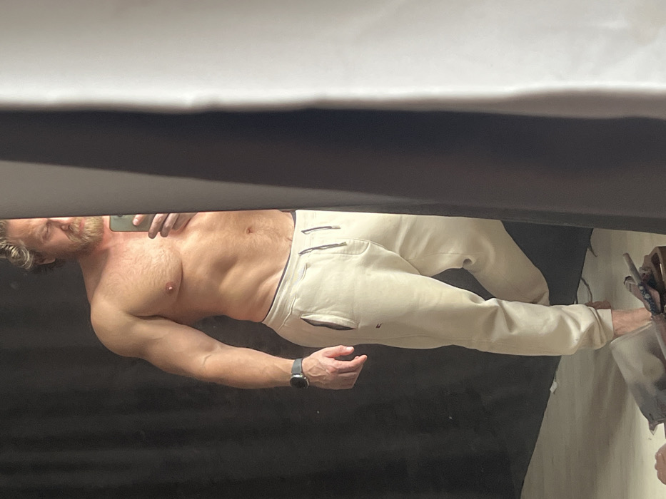 A muscular, shirtless male subject, visible from mid-thigh upwards, is depicted taking a mirror selfie with a dark smartphone held in his left hand. His left wrist bears a dark-strapped smartwatch. The subject has light-colored hair and a full beard; his face is partially obscured by the right edge of the mirror's reflection, revealing his right eye, nose, and bearded jawline. He wears light beige sweatpants featuring a visible drawstring and a small red and blue logo near the left pocket. His feet are bare. The background behind the subject, as seen in the mirror's reflection, is a uniform dark, likely black, surface, suggestive of a studio backdrop. To the right of the mirror's reflection, a portion of the room's actual interior is visible, consisting of a vertical dark grey surface bordering a lighter, possibly white, panel. On the floor, near the subject's bare feet, a grey metallic bar or pipe lies alongside a brown wooden object. The specific location within Saint Petersburg, Russia, cannot be determined from the visual information provided in the image.