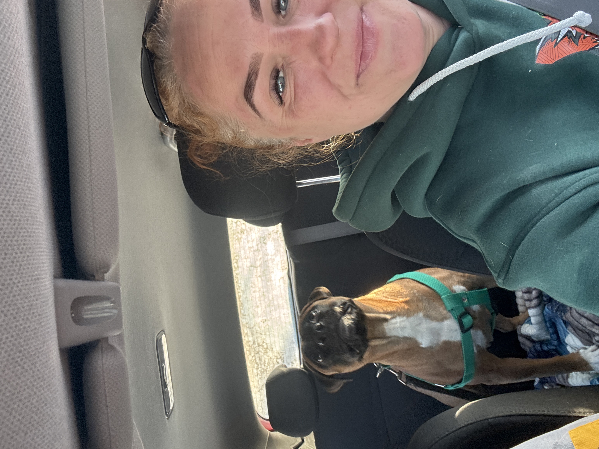 A woman is seated in the back seat of a car, possibly taking a selfie. She has fair skin, blue eyes, and light brown hair with darker eyebrows. She is wearing sunglasses perched on her head and a green hooded sweatshirt with a white drawstring. In the same back seat, a brown and white boxer dog is wearing a green harness. The car interior is gray. The location is assumed to be in the city of Chaikovsky, Russia but this cannot be determined from the image alone. The view through the car window is ambiguous.