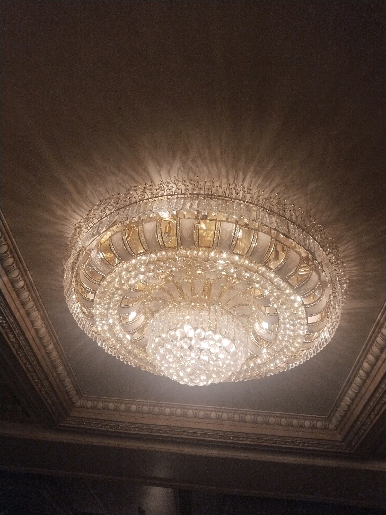 A large, multi-tiered crystal chandelier hangs from a dark, ornate ceiling in an indoor setting, likely a formal room or ballroom. The chandelier is illuminated, casting a warm glow and projecting intricate light patterns onto the ceiling and surrounding surfaces. The ceiling itself appears to be a dark, textured material with decorative molding around the edges. The chandelier is the primary focal point, with numerous clear, faceted crystals and gold-toned metalwork. The image is taken from a low angle, looking up at the fixture. There are no people or other objects visible in the frame. The specific location in Sinduadi, Indonesia is not discernible from the image itself.