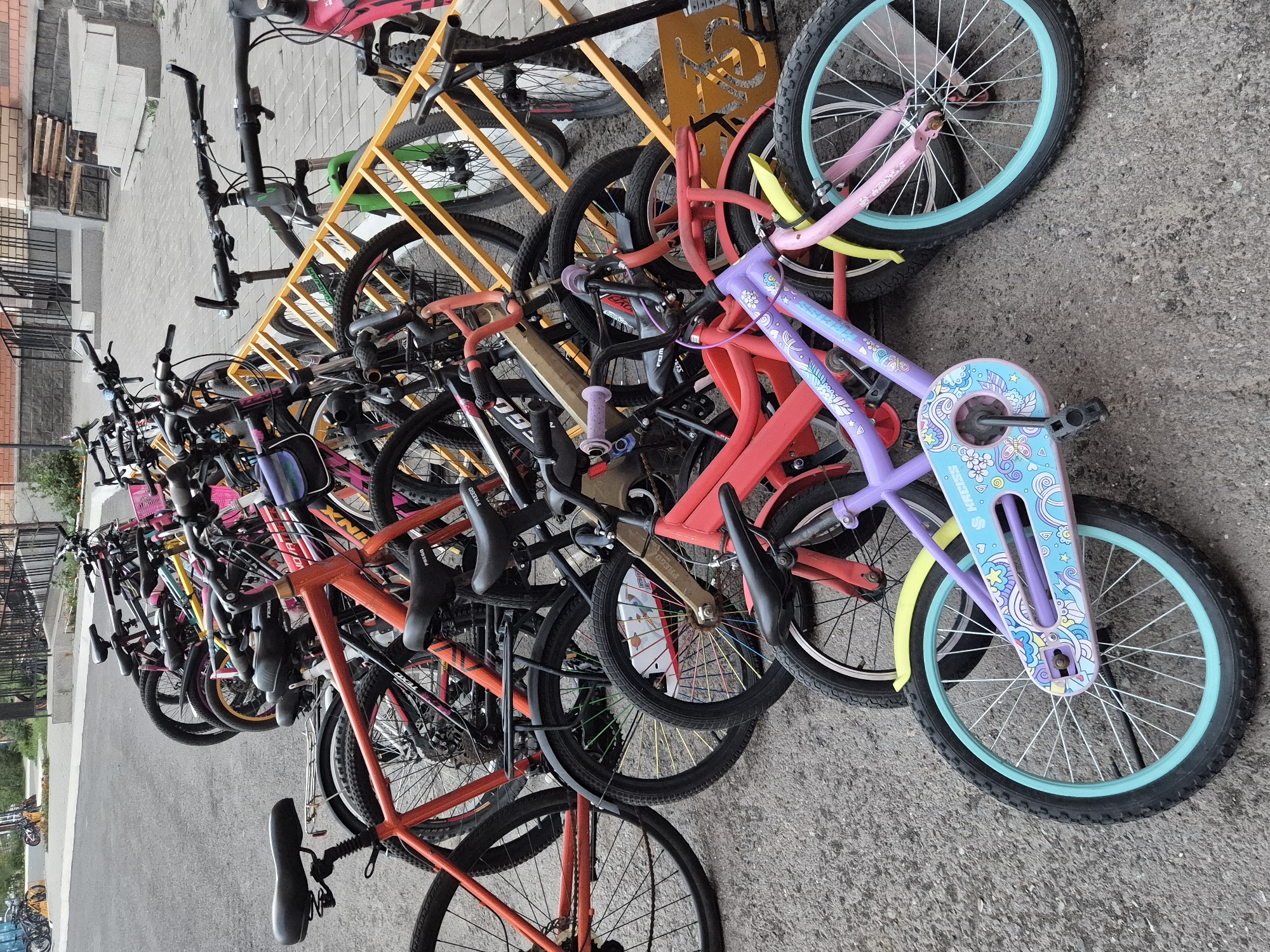 In Astana, Kazakhstan, a row of bicycles is parked at a rack in front of a building. The bicycles are of various colors and sizes, with some being adult-sized and others being children's bikes. The bicycles are leaning against a yellow metal rack. The ground is paved with asphalt. The building behind the bicycles has a brick facade and a set of stairs leading up to an entrance. The scene appears to be in a public area, possibly near a residential building or a school. The lighting suggests it is daytime.