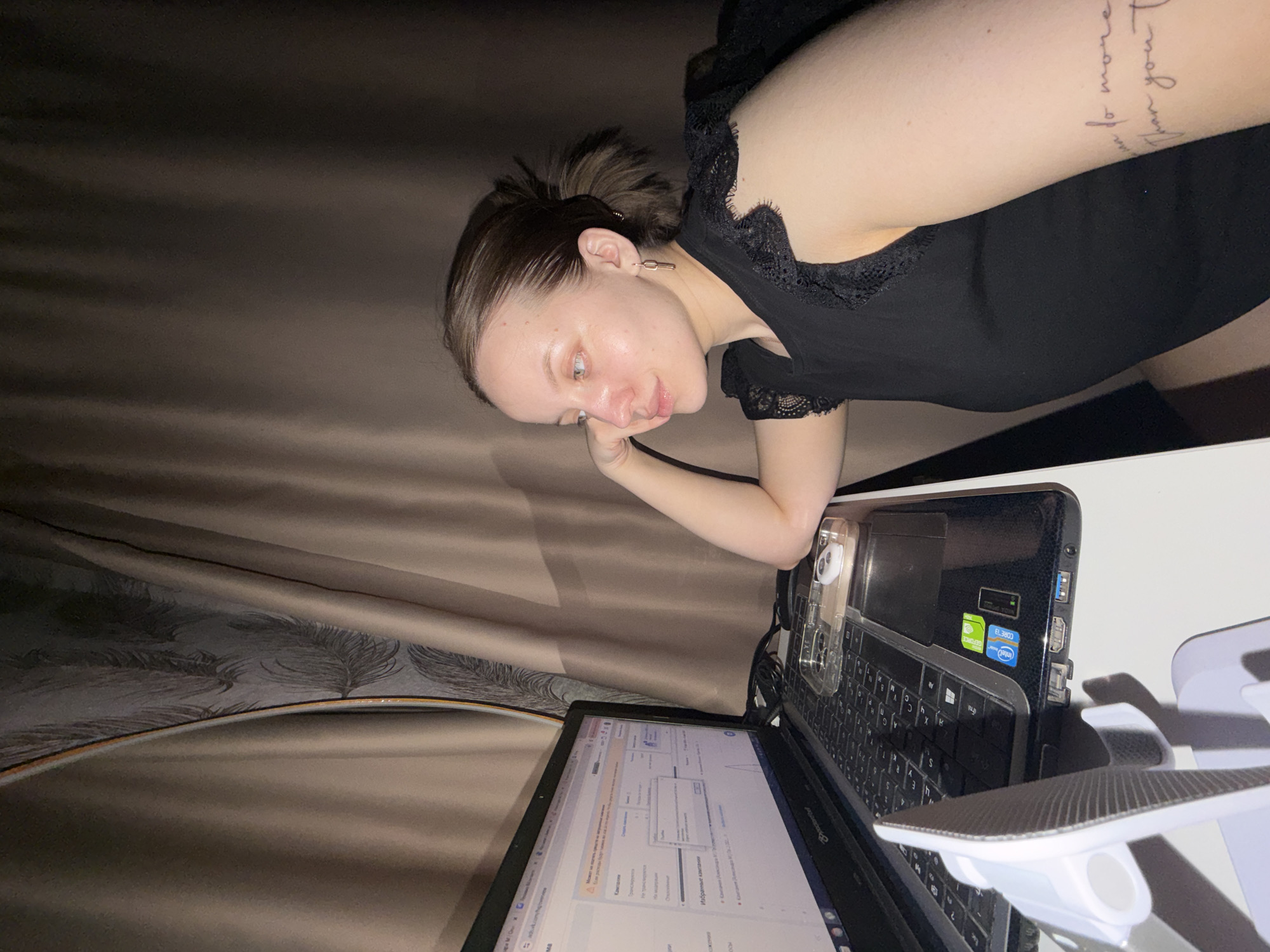 A woman with a tattoo on her upper arm is looking at a laptop screen. She is wearing a black top with lace trim. The laptop is sitting on a white surface, and there is a phone in a clear case next to it. There is also a white phone stand. The background appears to be a curtain. There is nothing to suggest the image was taken in Reshetnikovo, Russia.